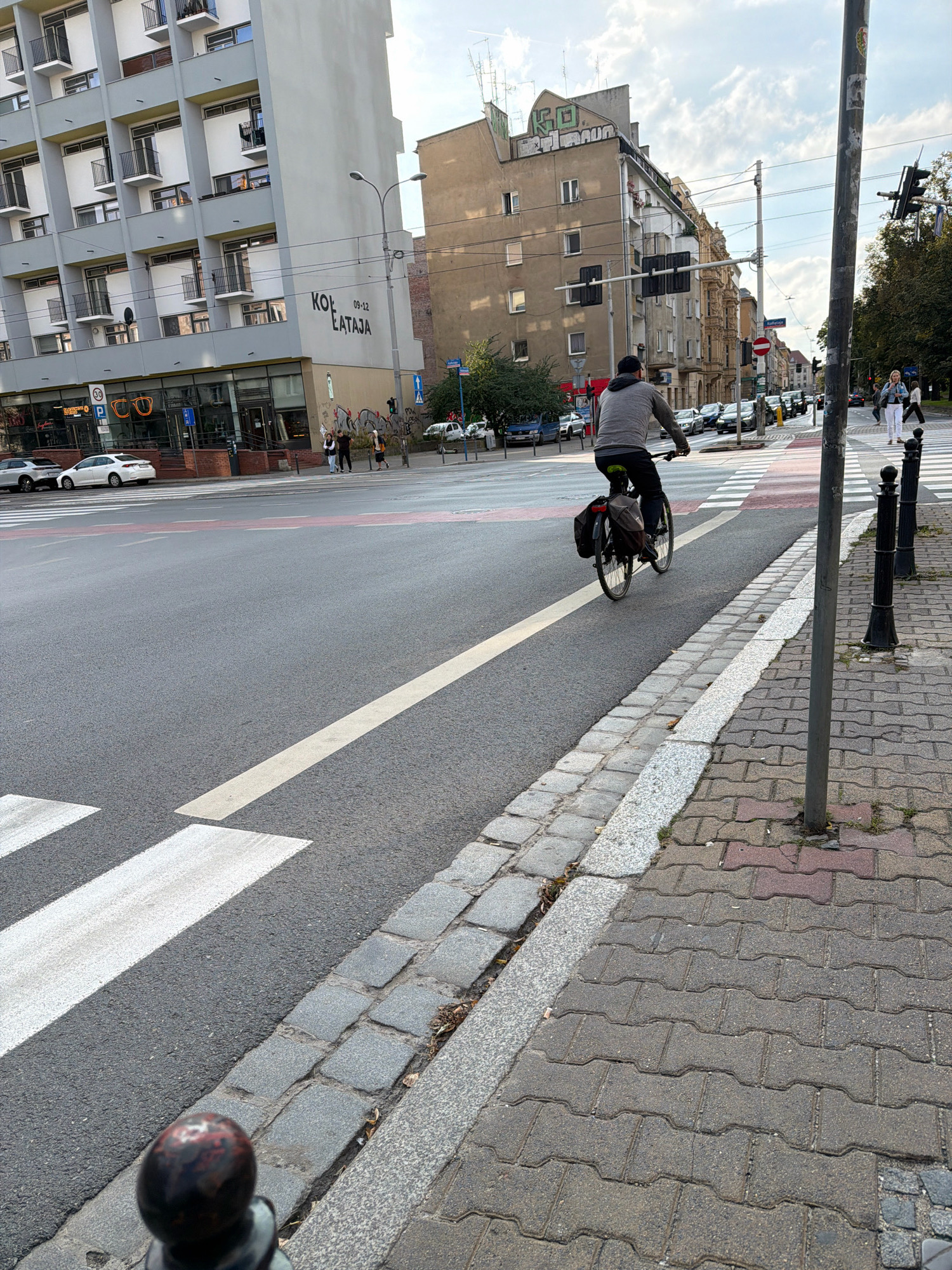 An outdoor, eye-level, full shot captures a street scene in Wrocław, Poland, on an overcast day. In the foreground, a grey-clad person is riding a bicycle with saddlebags on the rear rack. The cyclist is moving away from the camera, along a dedicated bike lane marked by a white line on the asphalt. The bike lane runs parallel to a pedestrian sidewalk paved with interlocking bricks and bordered by cobblestones.
The street features a pedestrian crossing with white stripes. Several cars are visible on the road, some parked and some in motion. To the left of the cyclist, a modern apartment building with numerous balconies and windows is prominent. A sign on the building reads "KOL EATAJA" and "09-12". Across the street, other buildings of varying architectural styles, including an older, darker building with graffiti on its upper facade, line the road. Traffic lights and street lamps are present. The sky is cloudy.