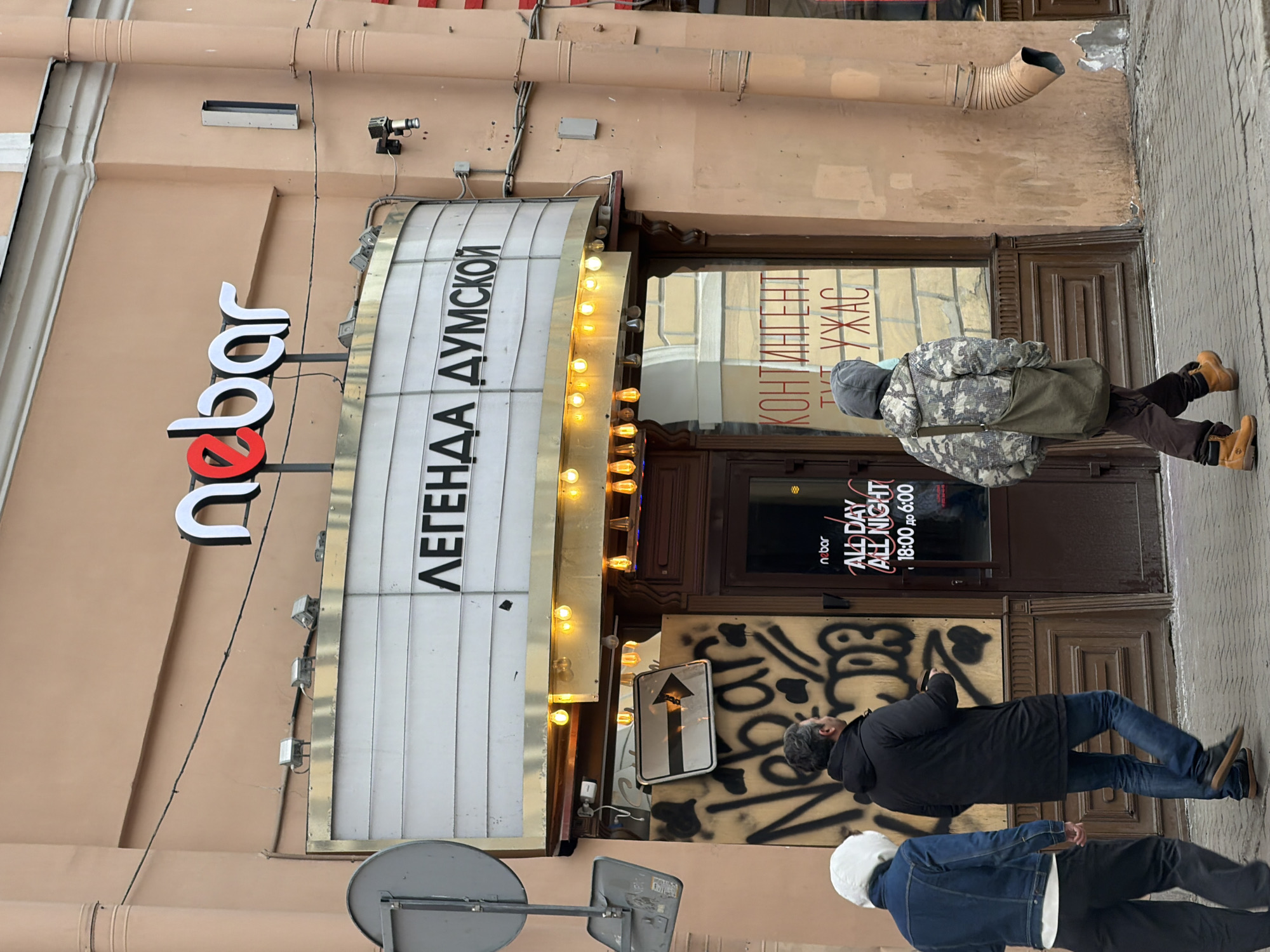 The image depicts an outdoor street scene in Saint Petersburg, Russia, specifically on Dumskaya Street, indicated by the marquee sign reading "ЛЕГЕНДА ДУМСКОЙ" (LEGEND OF DUMSKOY). The focal point is the entrance to a building with a light peach-colored facade, identified by a prominent sign as "nebar" (НЕБАР) in white and red lettering. Below this, a white marquee-style sign with horizontal slats displays the black Cyrillic text "ЛЕГЕНДА ДУМСКОЙ". A string of bare incandescent bulbs hangs directly below the marquee, illuminating the entrance area.
The entrance consists of a wooden-framed glass door, reflecting the interior signage that includes the "nebar" logo and operating hours: "ALL DAY ALL NIGHT 18:00 до 6:00" (18:00 to 06:00). Another reflection on the door shows red Cyrillic text "КОНТИНГЕНТ ТУТ УЖАС" (CONTINGENT HERE HORROR). To the left of the door, a section of the wall appears boarded up and is covered in black graffiti, which includes the word "Nebar". A white rectangular sign with a black right-pointing arrow is affixed over part of the graffiti. A grey corrugated downspout runs vertically down the right side of the building. Utility boxes and wiring are visible on the facade. The sky appears overcast.
Three individuals are walking on the paved sidewalk in front of the establishment, moving away from the viewer. The leftmost person wears a white hood or beanie, a blue denim jacket over a white garment, and dark pants. The middle individual, a male, is dressed in a dark hooded winter coat, blue jeans, and dark shoes; he is holding a dark object in his right hand. The rightmost person wears a camouflage-patterned hooded jacket, brown pants, and light brown work boots, carrying a dark green canvas shoulder bag. A light blue face mask is visible below this individual's chin. No direct interactions between the individuals or with the building are discernible.