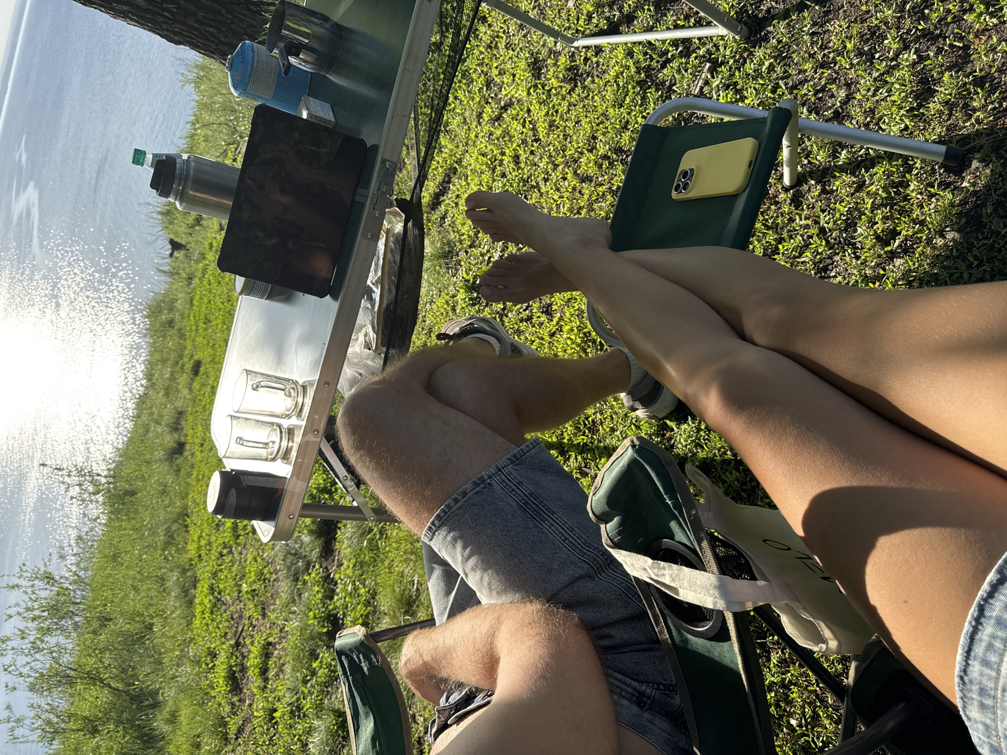 The image shows an outdoor setting with a metal folding table and two folding chairs positioned on grassy ground near a body of water reflecting sunlight. The table holds various items, including a water bottle, a camping stove, a tablet, and what appear to be glasses or mugs. The legs of two individuals are visible in the chairs. One person is wearing denim shorts and athletic shoes, while the other has bare feet and is wearing denim shorts. A yellow phone case is placed on one of the chairs. A tote bag is also visible on one of the chairs. The location appears to be a campsite near a lake or river. The city and state are unknown.