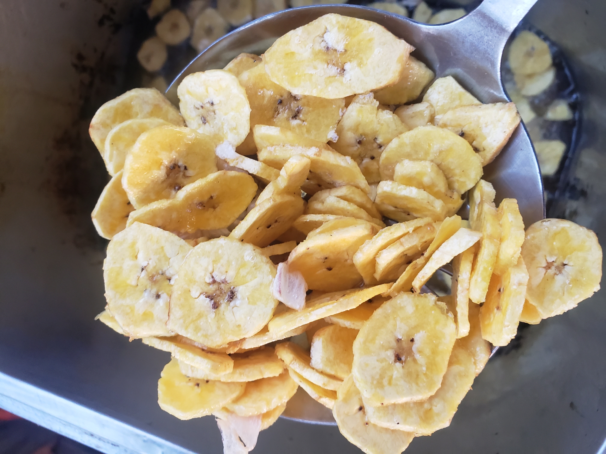 The image shows a close-up view of freshly fried plantain chips being lifted from a cooking vessel with a metal sieve. The plantain slices are round and golden brown, with some appearing slightly translucent from the frying process. Small pieces of white matter, possibly remnants of the plantain's skin or a seasoning, are visible among the chips. The background suggests a commercial kitchen setting, with a stainless steel counter to the left and what appears to be hot oil in the cooking vessel below the sieve, where more plantain slices are visible. The overall impression is one of food preparation in a bustling environment. The image is set in Narayi, Nigeria, a district known for its vibrant local markets and street food culture, where such delicacies are a popular offering.