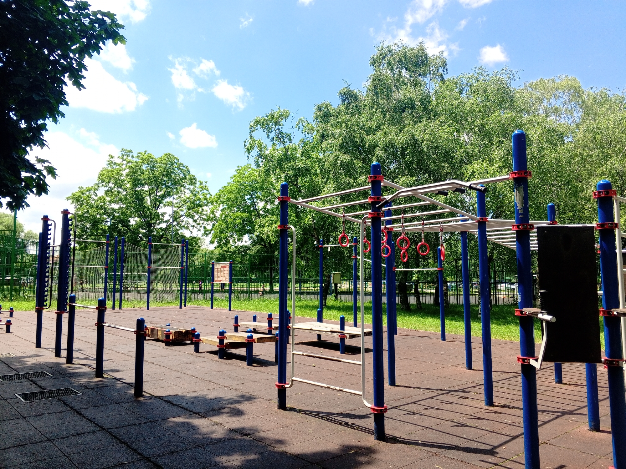 The image depicts an outdoor fitness park with various exercise equipment, situated in a park-like setting with trees and a blue sky with white clouds. The ground is covered with dark rubber tiles. In the foreground, there are several blue poles with red caps, supporting horizontal bars and rings. Some wooden benches are also visible. In the background, there are more exercise structures, a green fence, and lush green trees. There are no people visible in the image. The location is identified as Yessentukskaya, Russia.