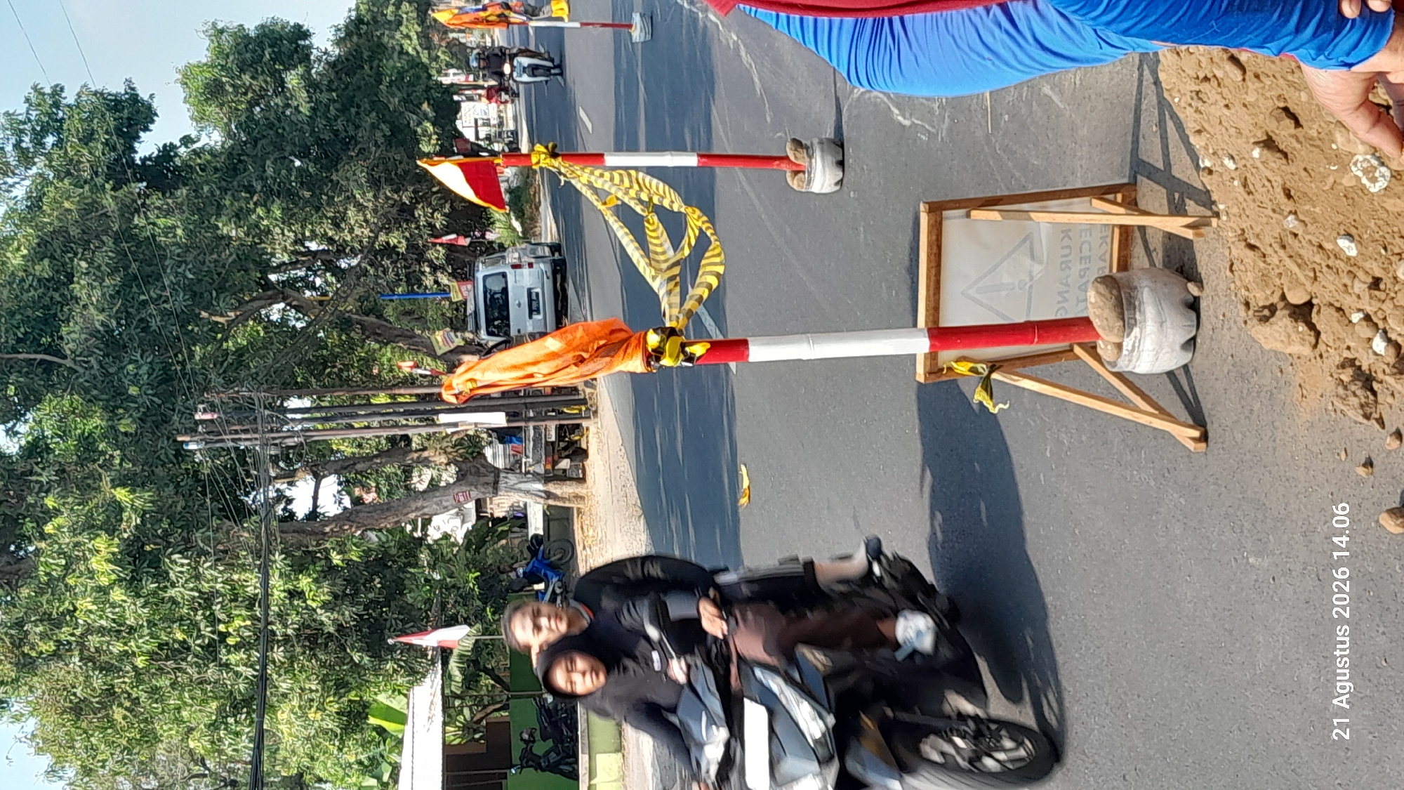 In the city of Takeran, Indonesia, on August 21, 2026, at 14:06, two individuals are riding a black motorcycle on an asphalt road. The motorcycle is moving from left to right in the frame, with the passenger in front wearing a black hijab and a black top, and the driver behind wearing a black long-sleeved shirt. The passenger is looking towards the left, and the driver is looking ahead. The road is bordered by trees on the left and right, with utility poles visible in the background. Several red and white striped posts with orange and yellow ribbons are present along the side of the road, suggesting potential roadwork or a temporary closure. A silver van is parked on the right side of the road in the background, and another motorcycle is visible further down the road. In the foreground, on the right side of the image, a person wearing a blue garment and red trim is partially visible, with their hand near a pile of dirt and rocks. A wooden easel with a sign is also positioned near the road. The weather appears to be sunny.