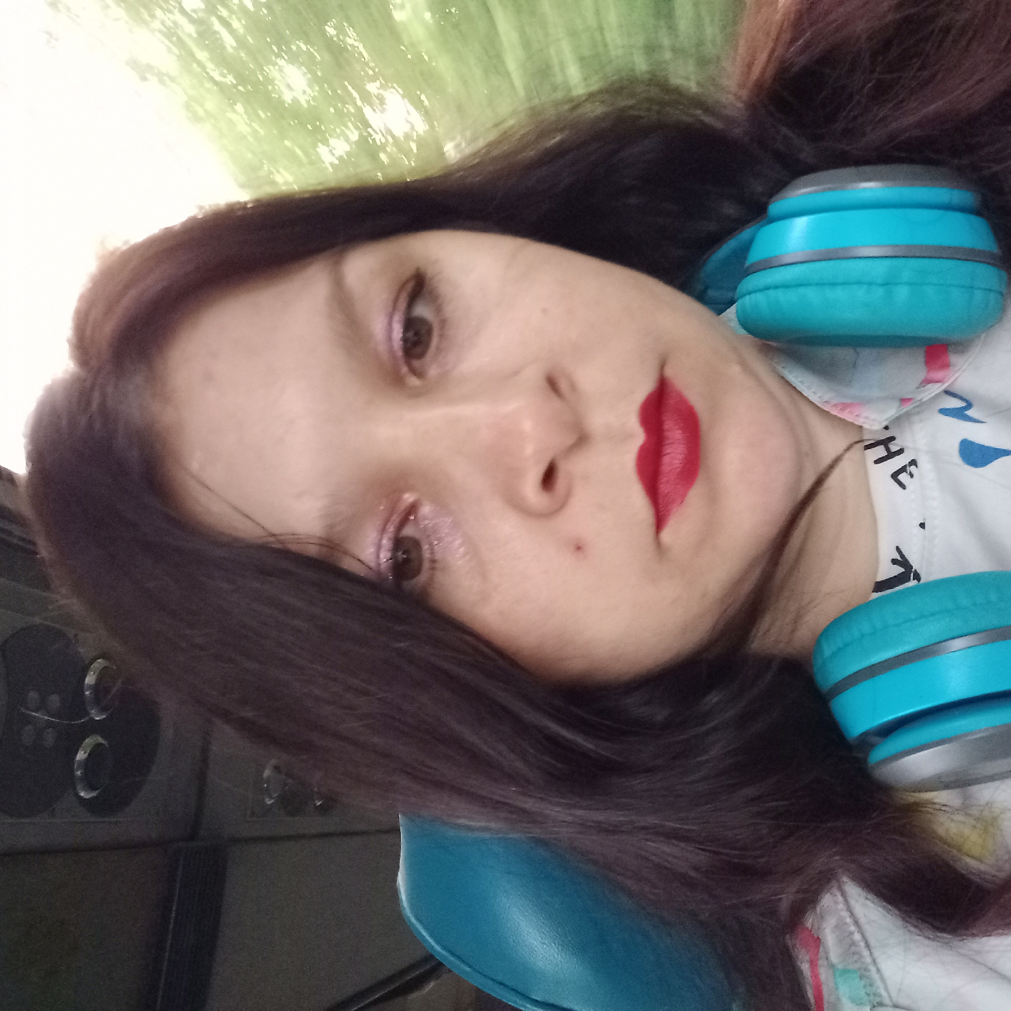 A selfie is taken by a woman with dark hair and red lipstick, wearing blue headphones. She is seated on a blue seat, likely on a form of public transportation, as indicated by the overhead compartments visible in the background. The environment appears to be outdoors, with a blurry green background suggesting foliage. The image is a close-up of the woman's face, with her headphones resting around her neck. The location is the city of Neopalymivka, Ukraine.