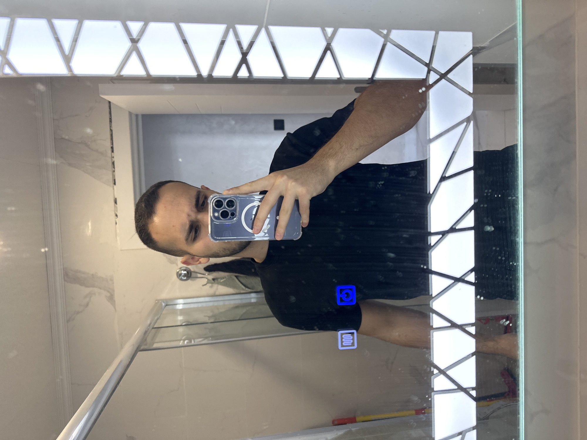 In Varna, Bulgaria, a person is taking a mirror selfie in a bathroom. The person is a male with short dark hair and is wearing a black t-shirt. He is holding up a smartphone to take the photo. The mirror has LED lighting around its edges and features two illuminated icons on its surface: one depicting wavy lines, likely indicating a defogger or heater, and another showing a power symbol. The bathroom environment is visible in the reflection, including a shower enclosure, tiled walls, and what appears to be a cleaning mop.