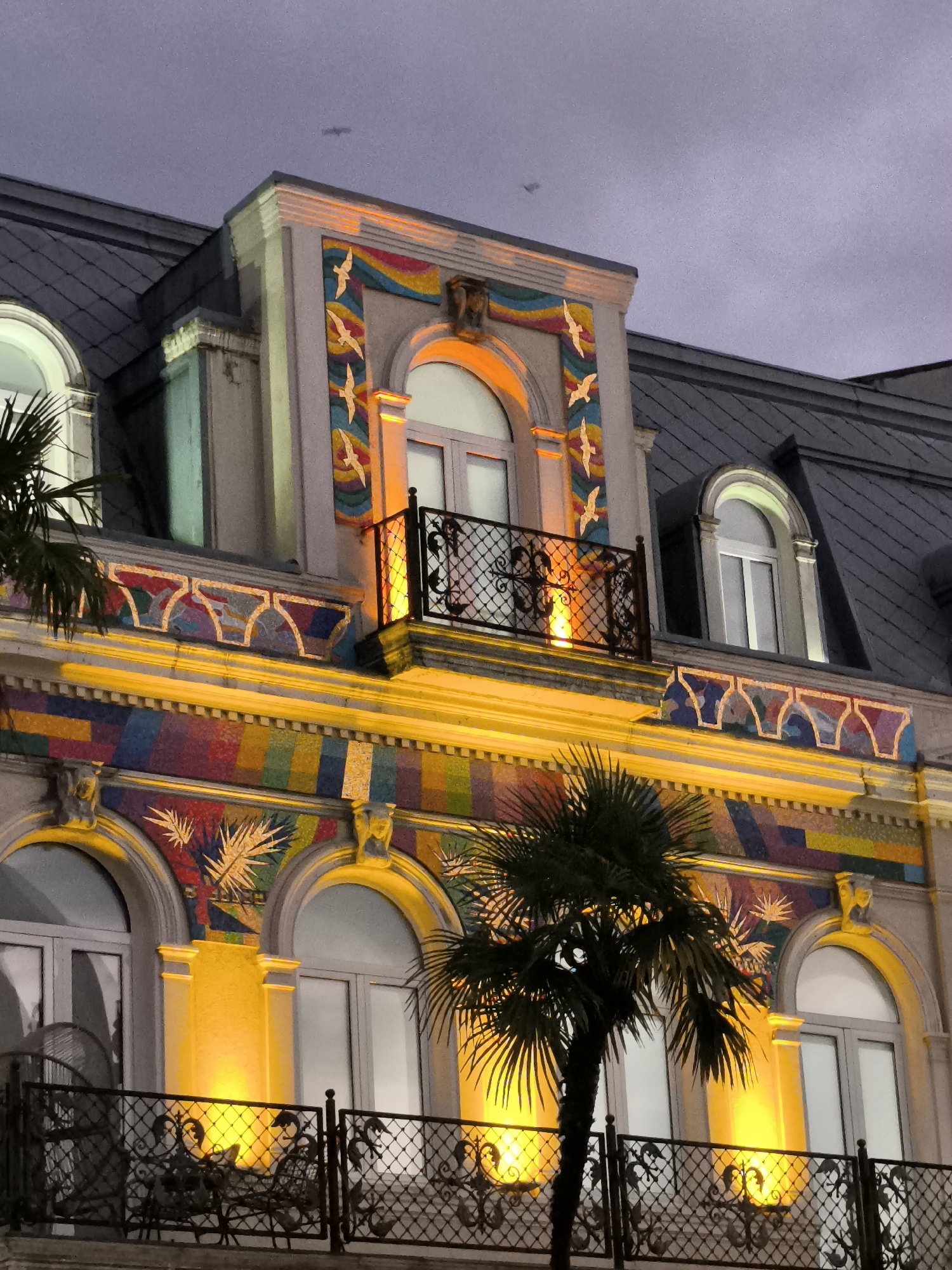 The image captures a close-up view of the colorful facade of a building, likely located in Batumi, Georgia, characterized by its Art Nouveau or eclectic architectural style. The building features a multi-colored mosaic, vibrant yellow lighting, and ornate balconies with intricate metal railings. Palm trees are visible in the foreground, suggesting a coastal or subtropical location. The sky is overcast and appears to be twilight or evening. There are no people or animals visible in the frame. The building's design incorporates arched windows, decorative moldings, and a series of stylized bird motifs in flight, rendered in mosaic form. The overall impression is one of opulence and artistic flair.