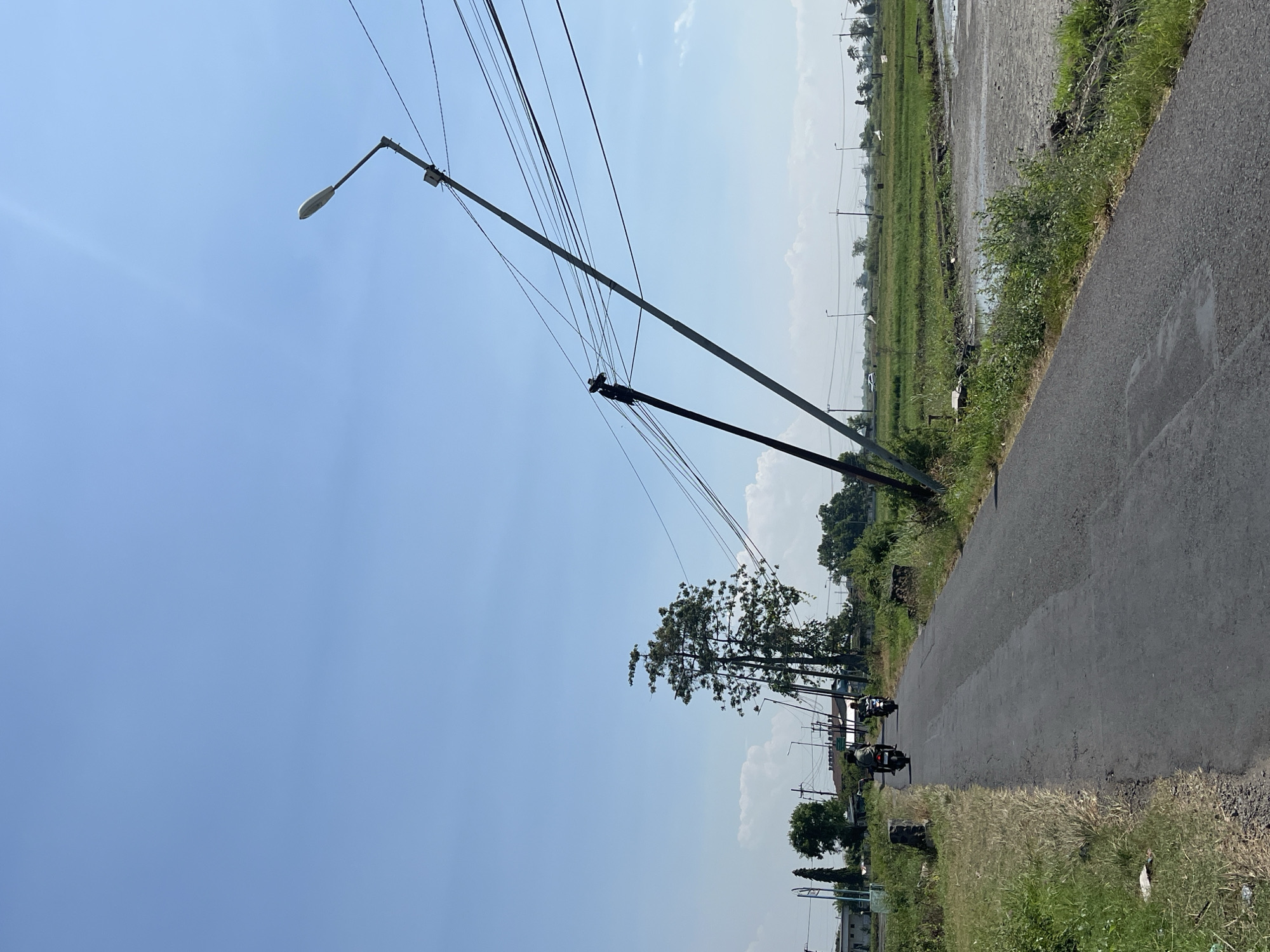 A rural road in Pesawahan, Indonesia, is depicted under a clear blue sky. Two people are riding motorcycles on the asphalt road. To the right of the road, a field of green crops stretches into the distance, bordered by more vegetation and a body of water. Two utility poles lean precariously, one significantly angled, with numerous electrical wires extending between them and across the scene. One of the poles is black and the other is grey. The scene is illuminated by bright sunlight.