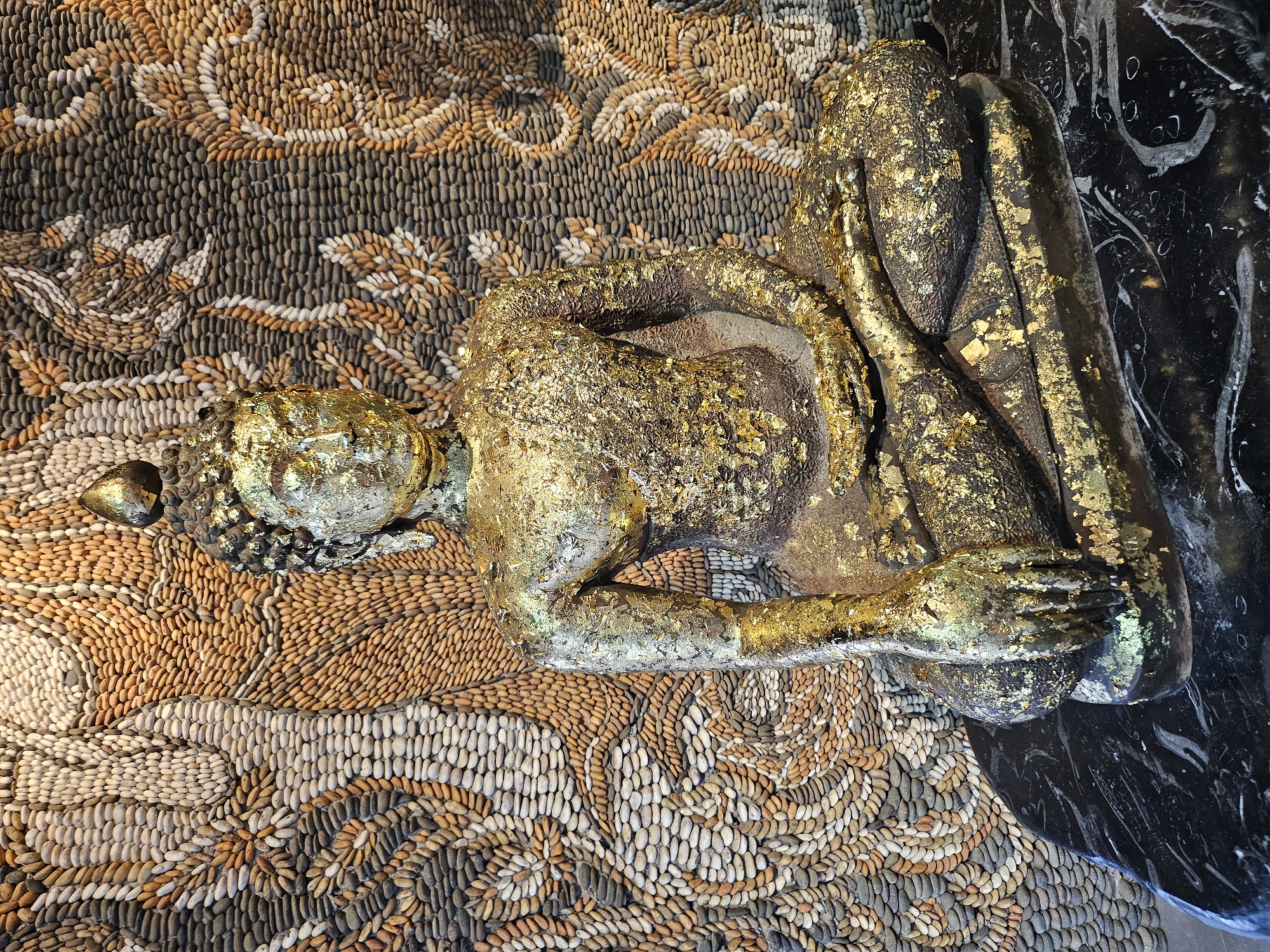 A seated Buddha statue, gilded with gold leaf, is positioned in front of a wall mosaic made of small, smooth pebbles. The mosaic depicts an abstract pattern in shades of brown, beige, and gray, resembling flowing organic forms. The Buddha statue itself is dark gray underneath the gold leaf, which is unevenly applied, with some areas of gold being more prominent than others. The statue has a serene expression and is seated in a lotus position. It rests on a dark, polished surface, possibly marble, with white veining. The background mosaic is highly detailed and covers the entire wall behind the statue. The lighting is soft, casting subtle shadows. This scene is located at the Sanctuary of Truth in Pattaya, Thailand, which is near Na Chom Thian.