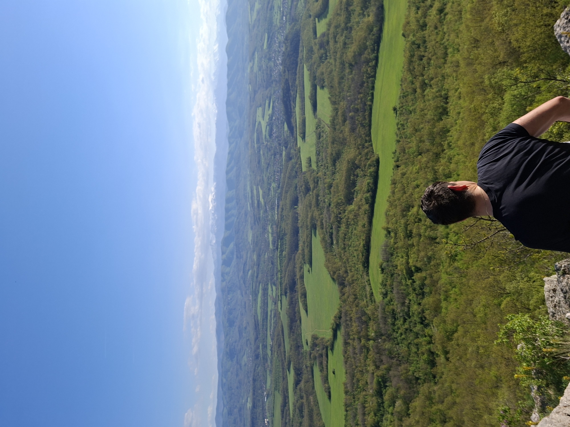 The image captures a scenic view from what appears to be a high vantage point in Zdravkovets, Bulgaria. The foreground features a person, partially visible on the right, wearing a black shirt and standing near a rocky outcrop. The middle ground showcases a landscape with alternating patches of green fields and dense forest areas. In the background, a mountain range stretches across the horizon under a bright blue sky with some clouds.