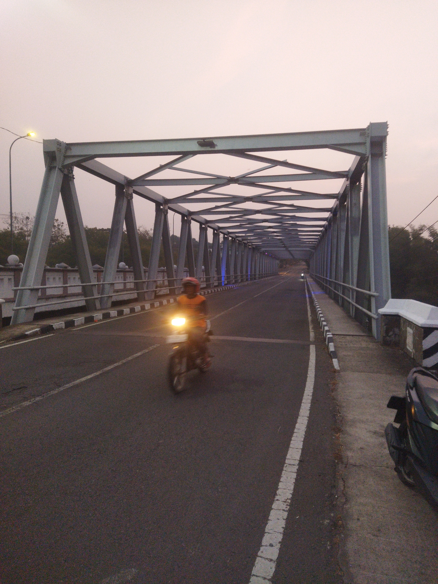 A lone motorcyclist wearing an orange jacket and a helmet rides down the center lane of a bridge in Wonogiri, Indonesia. The bridge features a steel truss structure and a concrete road. The sky is overcast and a dull grey, suggesting either dawn or dusk. The road is marked with white lines, and the bridge is lined with a low-rise white barrier topped with decorative spheres. To the right of the road, a portion of a parked dark-colored motorcycle is visible. The overall atmosphere is quiet and slightly eerie, typical of early morning or late evening in a less populated area.