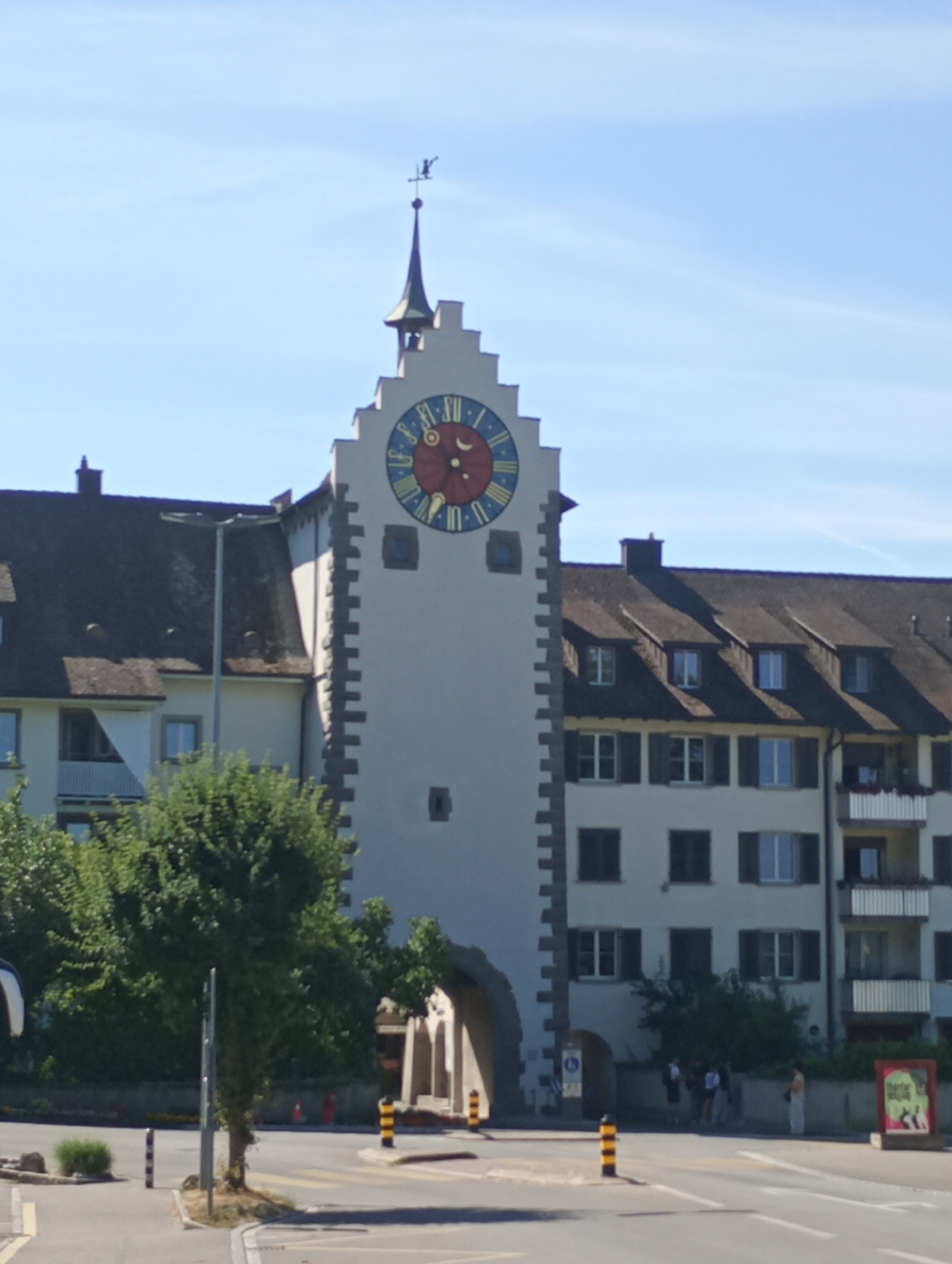 A high-angle, full shot captures a white clock tower with a pointed roof and a weather vane, set against a clear blue sky. The tower is adorned with a large, colorful clock face showing the time as approximately 3:50. Below the clock, a dark arched gateway opens into a shadowed area, where several people stand. The clock tower is flanked by buildings with dark roofs and multiple windows. To the left of the tower, a section of a lighter-colored building with a dark, sloped roof is visible, featuring a balcony with a white railing. To the right, a multi-story residential building with several balconies is present. In the foreground, a street intersection is shown, with a pedestrian crossing marked by white lines. Yellow and black bollards line the intersection, and a street lamp stands near the center of the frame. Trees and shrubs are scattered throughout the scene. The overall scene depicts a street in the historic town of Stein am Rhein, Switzerland.