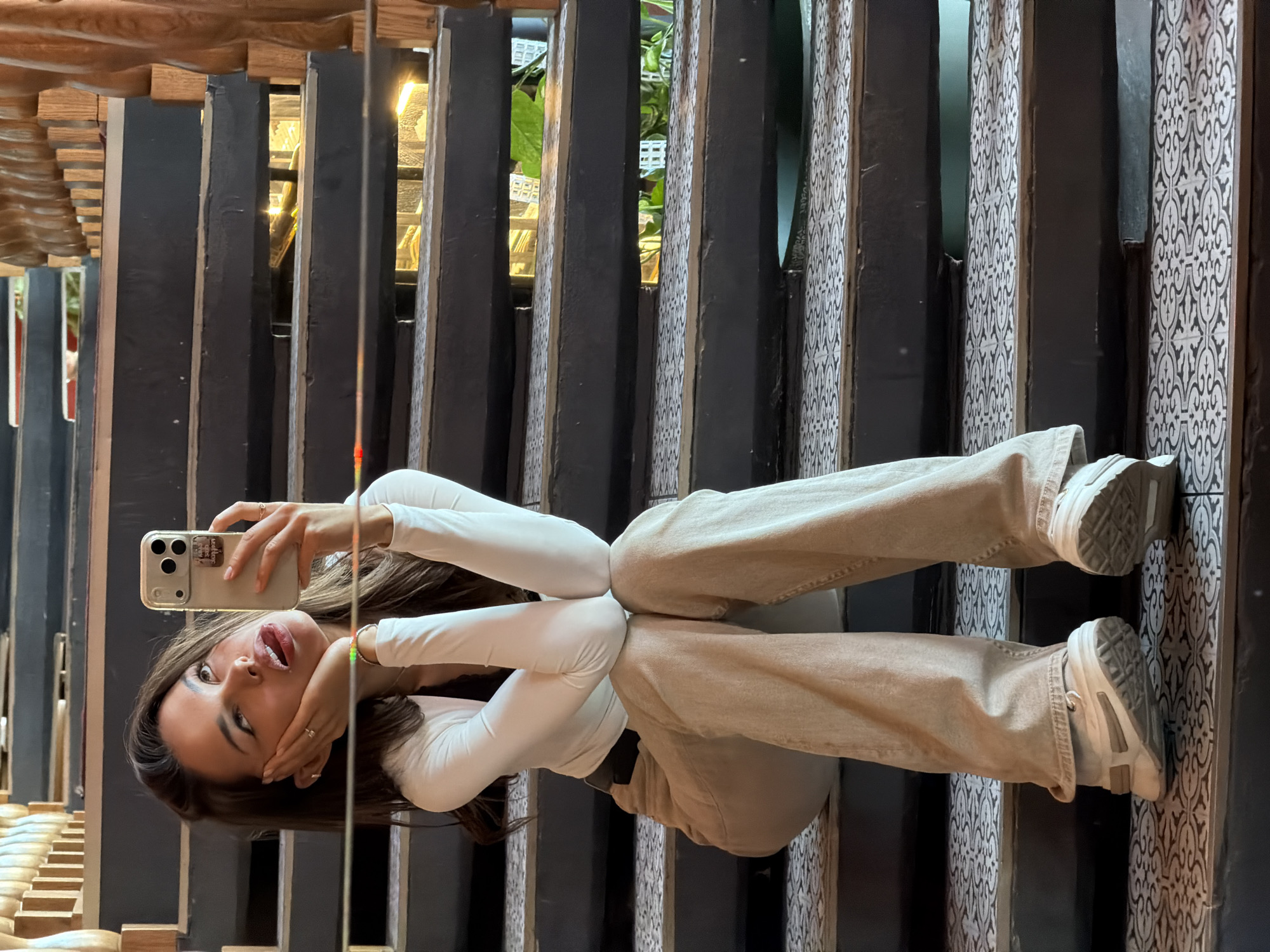 A young woman is taking a selfie while sitting on a black staircase with patterned tiles. She is wearing a white long-sleeved shirt and beige pants. Her left hand is supporting her head, and her tongue is sticking out. She is wearing white sneakers and several rings. The staircase has wooden railings and is illuminated by warm light. The background appears to be an indoor space with greenery visible through the gaps in the stairs. The woman's reflection is visible on the polished surface of a staircase step. The setting is not specified to be in Saint Petersburg, Russia.