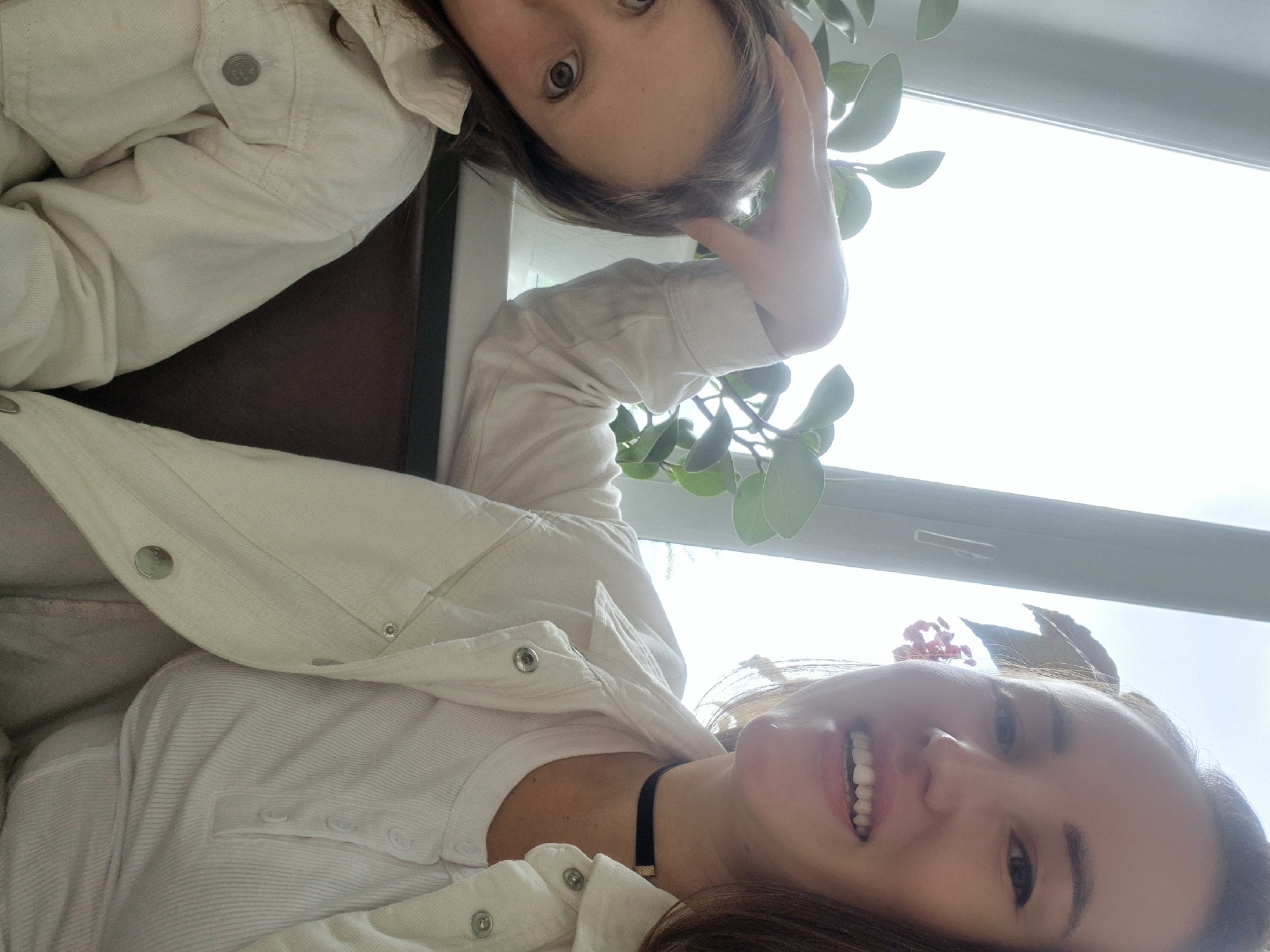 Two young women are indoors, positioned in a way that suggests they are taking a selfie. The woman in the foreground smiles broadly, revealing her teeth. She wears a white button-down shirt with a denim jacket over it and a black choker. The other woman is in the background, looking directly at the camera with her hand near her head. She is also wearing a white denim jacket. A plant with round leaves is visible between them, and a window with bright light coming through can be seen behind the plant. The location is not specified.