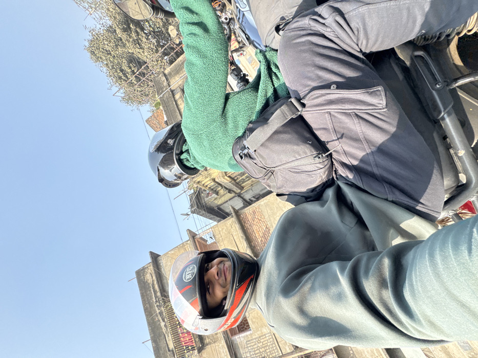 A bright, daytime photograph captures two individuals on a motorcycle in an urban environment, likely Kolkata, India. The image is taken from a low angle, looking slightly upwards, and appears rotated, with the horizon diagonal.
The person in the foreground, presumed to be the rider, wears a light grey or light green hooded sweatshirt and a distinctive black, red, and white motorcycle helmet. The helmet's visor is raised, revealing their face, which shows dark hair and a neutral to subtly smiling expression as they look towards the camera.
Behind them, the pillion rider is seen wearing a dark green, fuzzy-textured jacket and a glossy black full-face helmet with the visor lowered, obscuring their face. This individual is also dressed in dark grey or black cargo-style pants, featuring prominent pockets on the thighs. A black sling bag or fanny pack is worn across their chest or waist.
The motorcycle itself is partially visible, showing elements of its frame, seat, and handlebars. The activity appears to be riding or pausing during a ride, with the rider acknowledging the camera.
The background features several multi-story buildings constructed from brick and concrete, some showing signs of wear and exposed electrical wiring, characteristic of many urban areas in India. Bare tree branches are visible in the upper left. The sky is a clear, bright blue, indicating sunny weather. The strong sunlight casts distinct shadows, suggesting it is midday. No discernible text is clearly visible on the helmets, clothing, or in the background.
