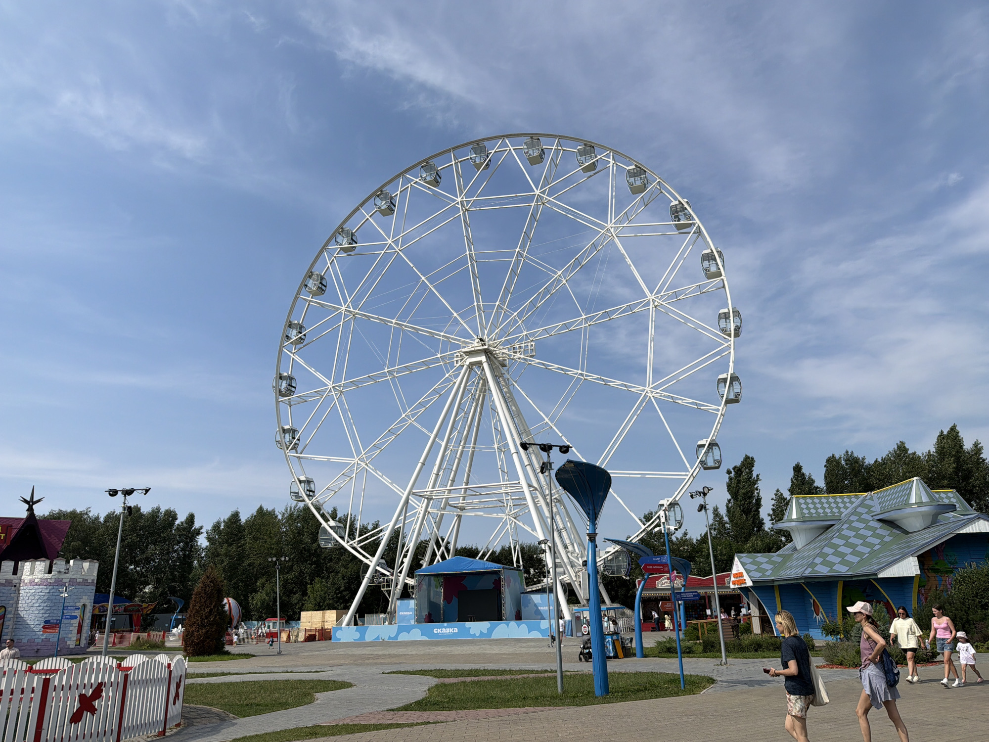 In Khimki, Russia, a large white Ferris wheel stands prominently against a bright blue sky with scattered clouds. The Ferris wheel is the central focus of the image, with its intricate white framework and numerous enclosed gondolas. In the foreground, a paved walkway curves through a park-like setting. Several people are walking on the path; on the right, three women and a child are walking towards the right side of the frame, with two women in the foreground and the other two following behind. A white picket fence with red accents runs along the left side of the walkway. Behind the fence, a whimsical, castle-like structure painted in blue and white with a dark purple roof is visible. Further back, trees form a dense green backdrop. To the right of the Ferris wheel, several colorful buildings with distinctive patterned roofs, likely part of an amusement park, are situated. A stage with a blue canopy and colorful graphics is set up in front of the Ferris wheel. The overall scene depicts a sunny day at an outdoor amusement park.