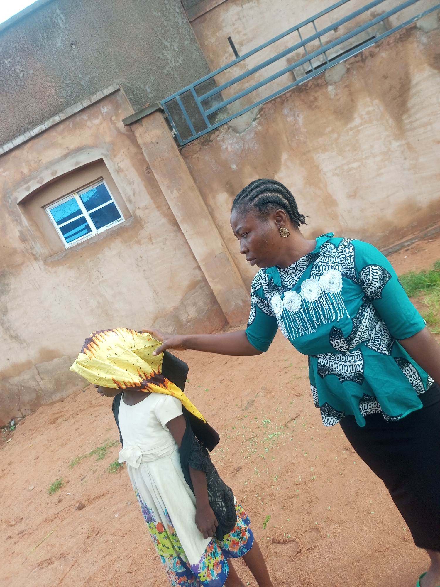In Tempure, Nigeria, a woman with braided hair, wearing a green and white patterned top and a black skirt, stands outdoors and places her hand on the head of a child. The child is wearing a white dress with a patterned skirt and a yellow headscarf that covers their face. They are also carrying a black bag. The background shows a building with a textured, brown-colored wall and a window with blue panes. The ground is covered in sand with scattered leaves. A metal railing is visible on the building's exterior.