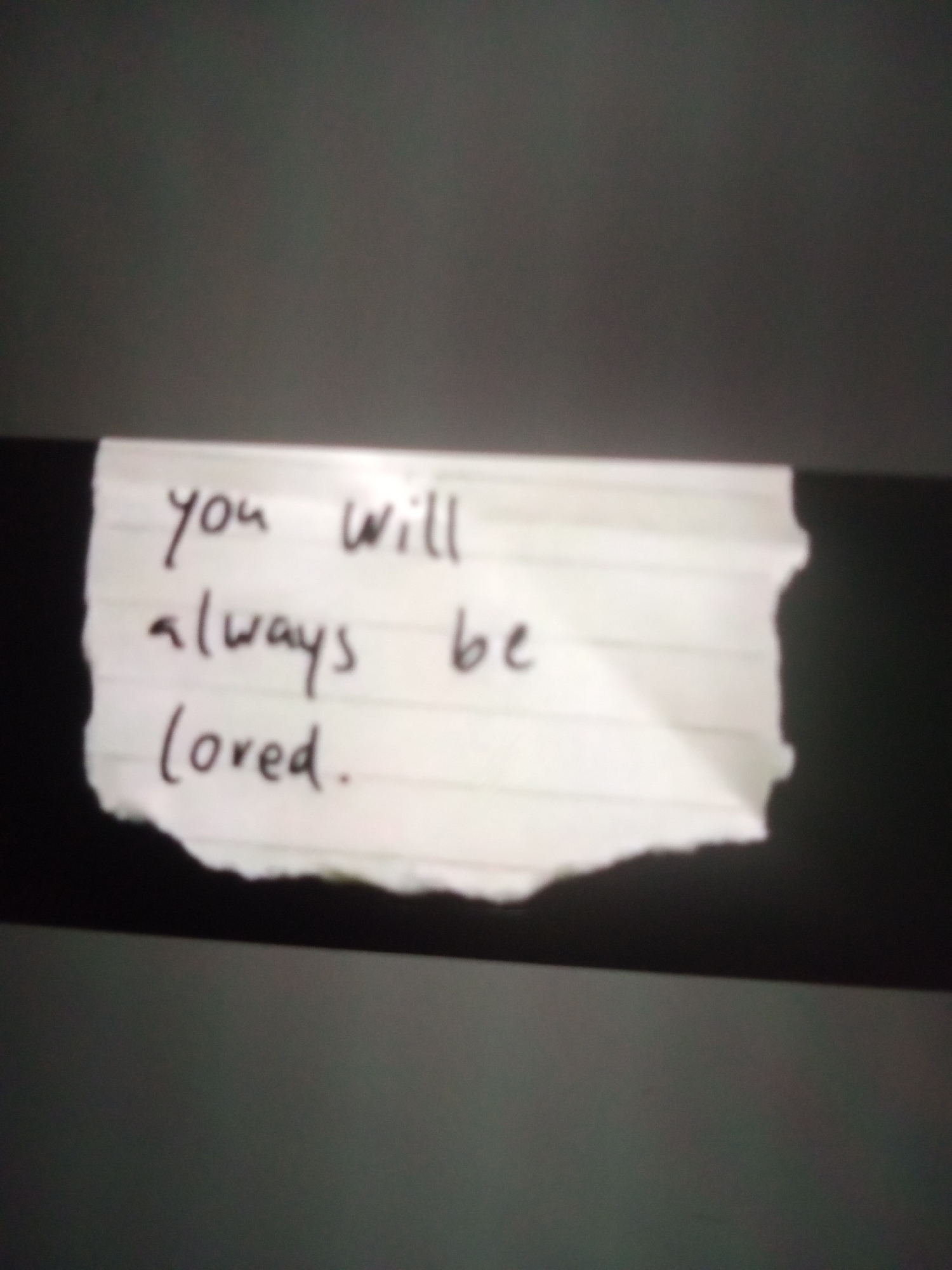 The image shows a torn piece of lined paper with handwritten text. The text reads "you will always be loved." The paper is on a dark background, with some of the paper torn away, revealing more of the dark background. There is no discernible location in the city of Jalingo, Nigeria, as the image is a close-up of the paper and its surroundings are not visible.