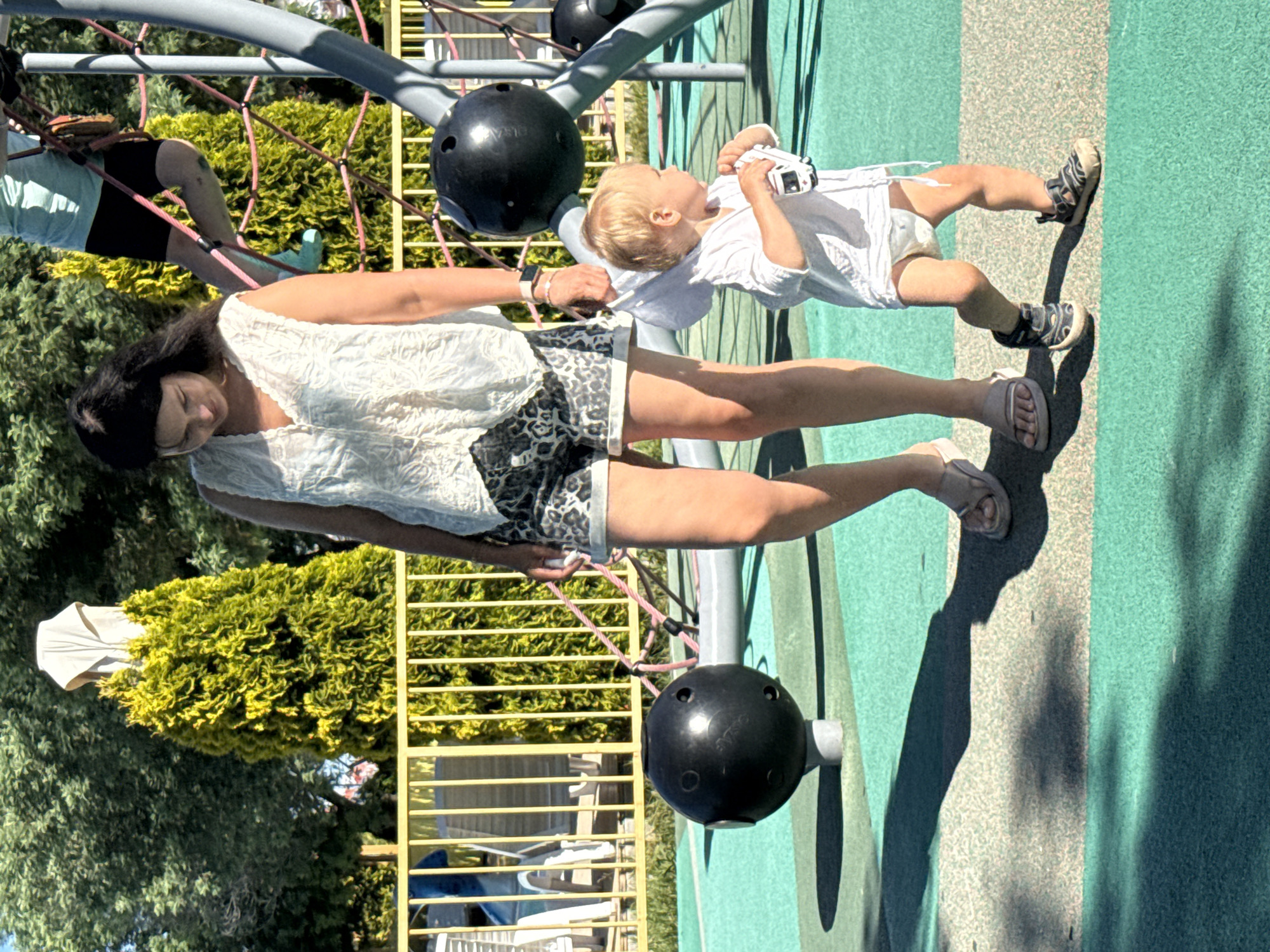 A woman and a toddler are standing on a playground in Sochi, Russia. The woman is wearing a white sleeveless top and animal-print shorts. The toddler, dressed in a white shirt and diaper, is holding a toy car. A jungle gym structure is visible in the background, along with trees and a yellow fence. The playground surface is a mosaic of green and light-colored rubberized material, with shadows cast by the sun. The woman appears to be holding the toddler's hand, as they are close together and both looking in the same direction.