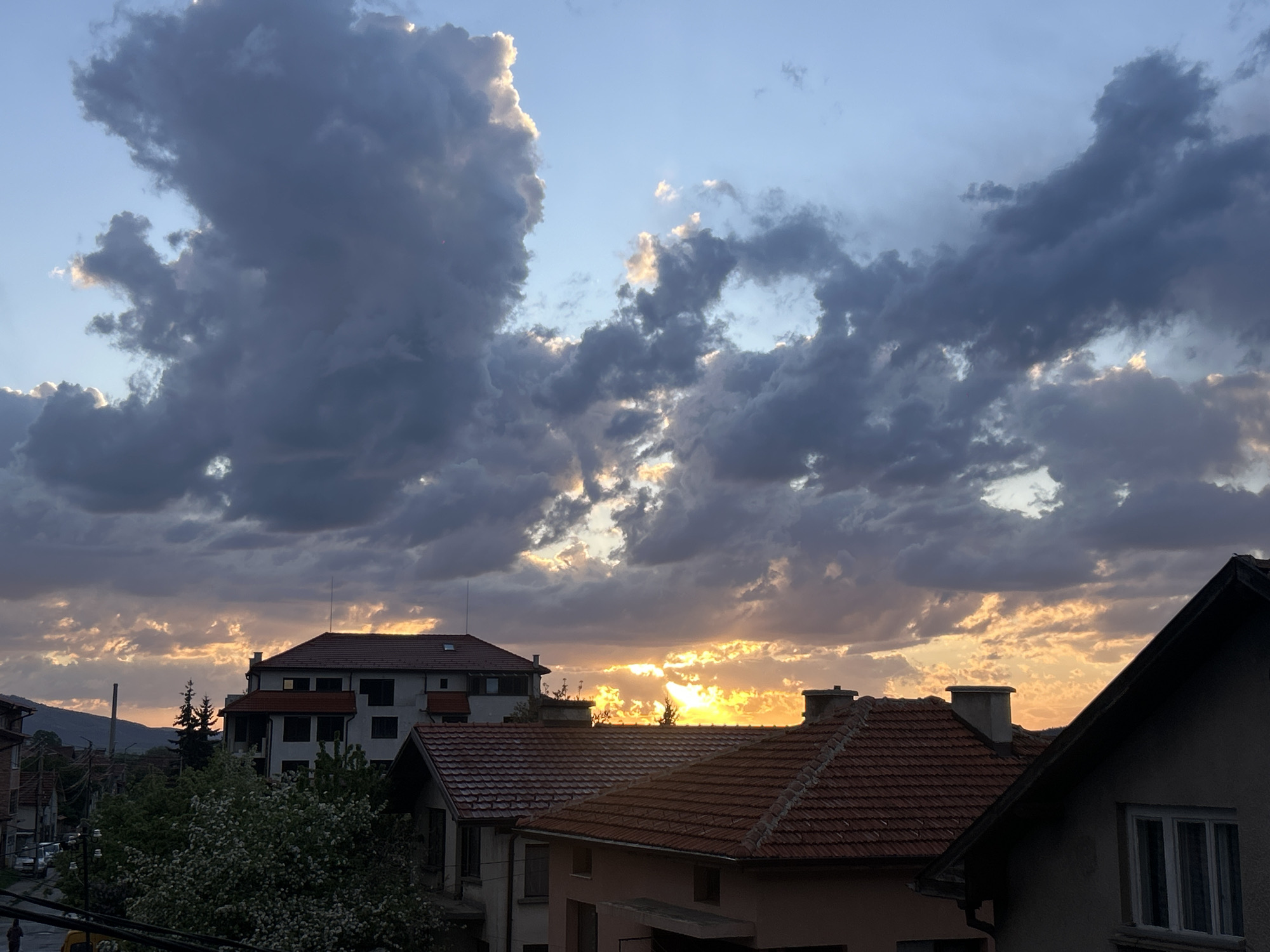 The image captures a twilight scene over an urban area. In the foreground, the roofs of several buildings with orange-tiled roofs are visible. A light-colored building stands further back with a red-tiled roof and multiple windows. Below it, greenery is present, including flowering trees.
The sky is filled with dense, dark clouds illuminated by the setting sun, which creates a vibrant yellow and orange glow. Some light streams through the clouds. A street is partially visible to the left with parked cars. In the distance, a hill or small mountain is silhouetted against the sky. The city is referred to as Unknown, Unknown.
