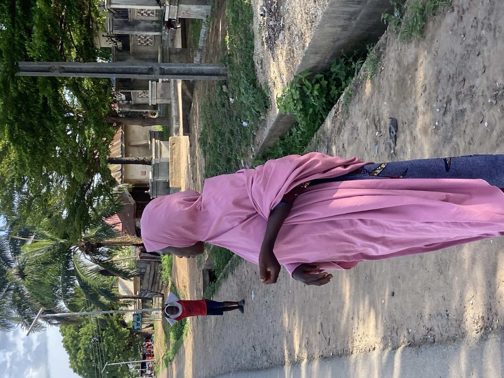 The image depicts a street scene in Mararaba, Nigeria. A woman wearing a pink hijab and a blue patterned garment is walking along a dirt road. In the background, a child in a red shirt and shorts is walking in the opposite direction. There are trees and buildings visible in the distance. The road is lined with concrete curbs and overgrown vegetation.