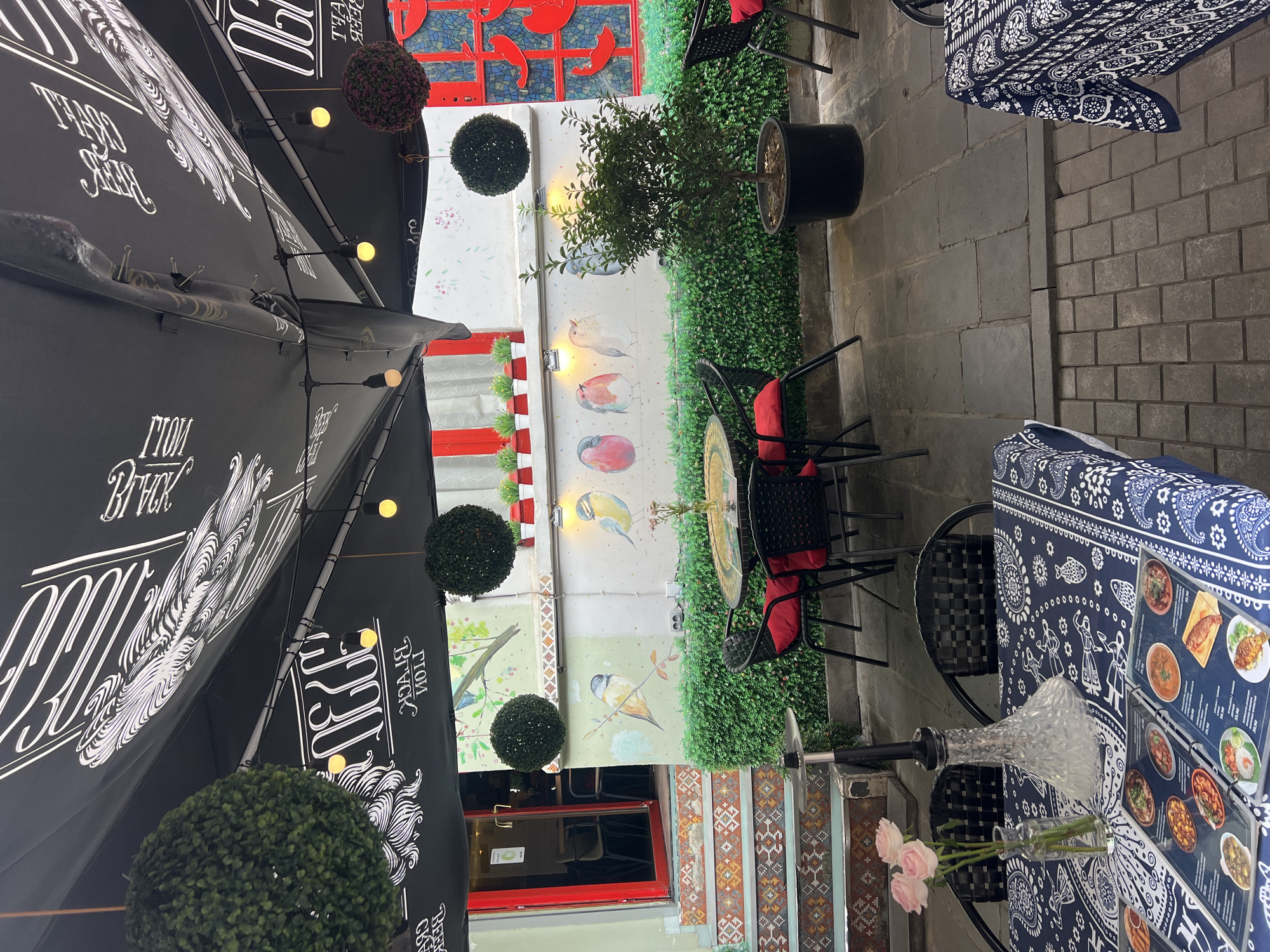 The image depicts an outdoor seating area of a cafe in Tbilisi, Georgia. The area is partially covered by a large black umbrella with white lettering. There are several tables set up, some with blue and white patterned tablecloths. On one table, a hookah, a vase with pink roses, and an open menu are visible. The walls are decorated with paintings of birds. In the background, there are red-framed windows and a doorway leading into the cafe. Lush green shrubbery lines the back of the seating area. The ground is paved with stone tiles.