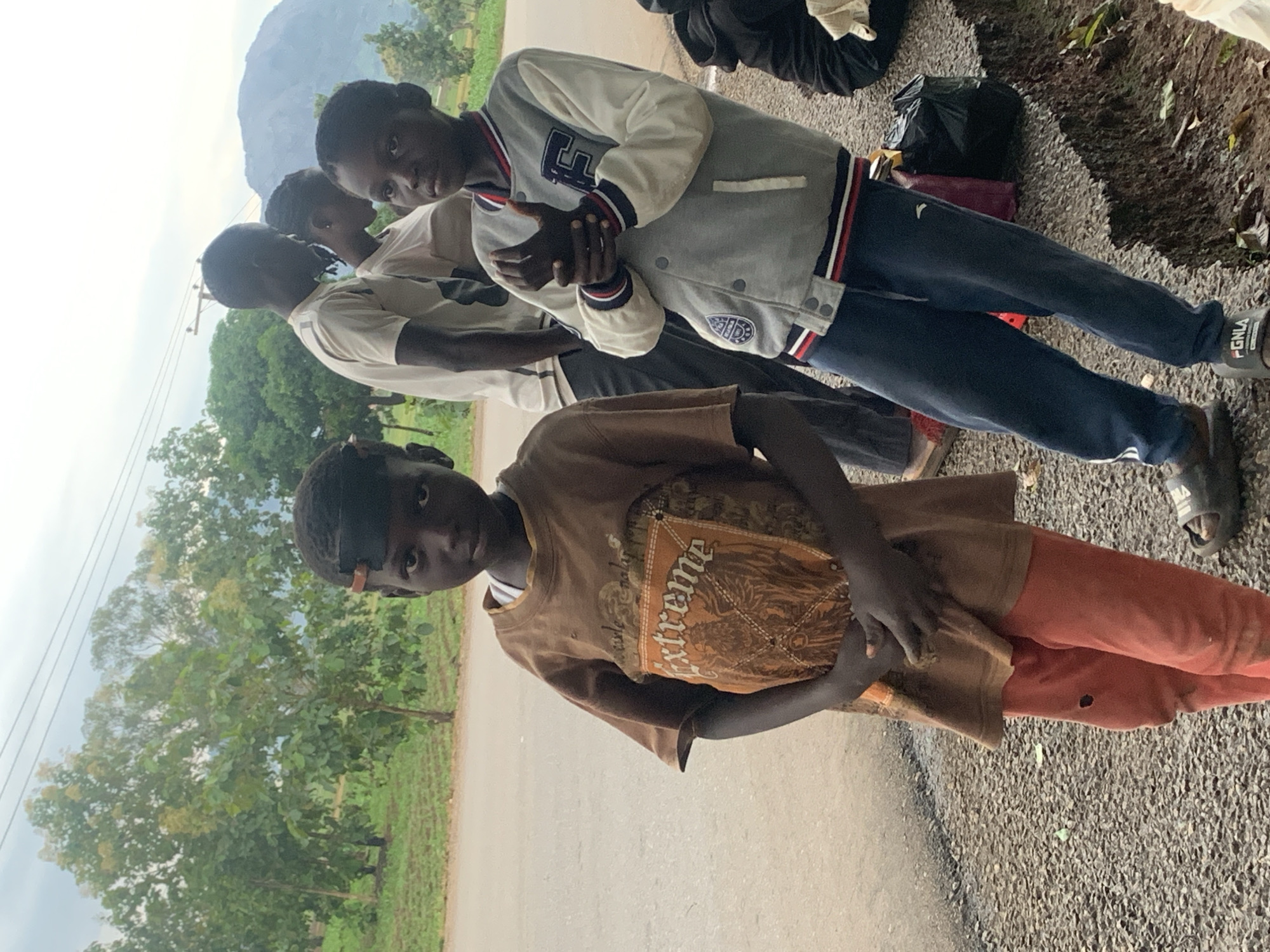 In the city of Agban, Nigeria, a group of young people stand by a road. The foreground features two boys. The boy on the left, with a dark headband, wears a brown t-shirt with the word "Extreme" printed in large letters and orange pants with a hole in the left knee. His hands are clasped in front of him. The boy to his right wears a grey and white jacket with blue and red trim, dark blue sweatpants, and dark sandals. He is looking down and holding his hands clasped together. Behind them, two other individuals, a young man and a young woman, are partially visible, also by the roadside. The environment appears to be a rural or semi-urban area with trees and a mountain in the background. The ground is covered with gravel and some vegetation. The sky is overcast.
