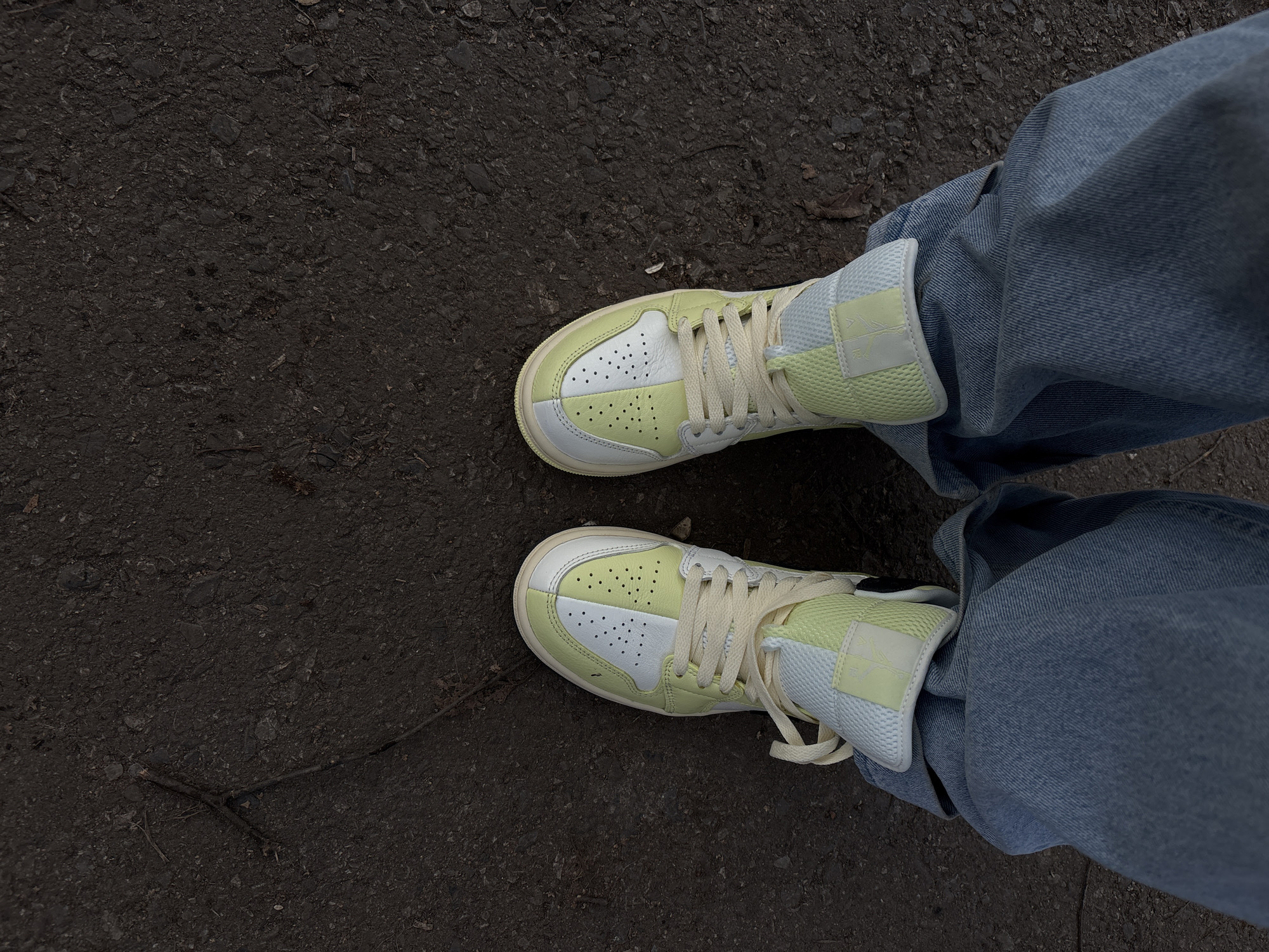 An overhead shot shows a person standing on dark asphalt, wearing light wash denim jeans and light green and white sneakers. The location appears to be outdoors, based on the dark asphalt surface. No other people, objects, or locations are explicitly identifiable in the image. No interaction can be assumed. The image does not show any details that would confirm Ulyanovsk, Russia.