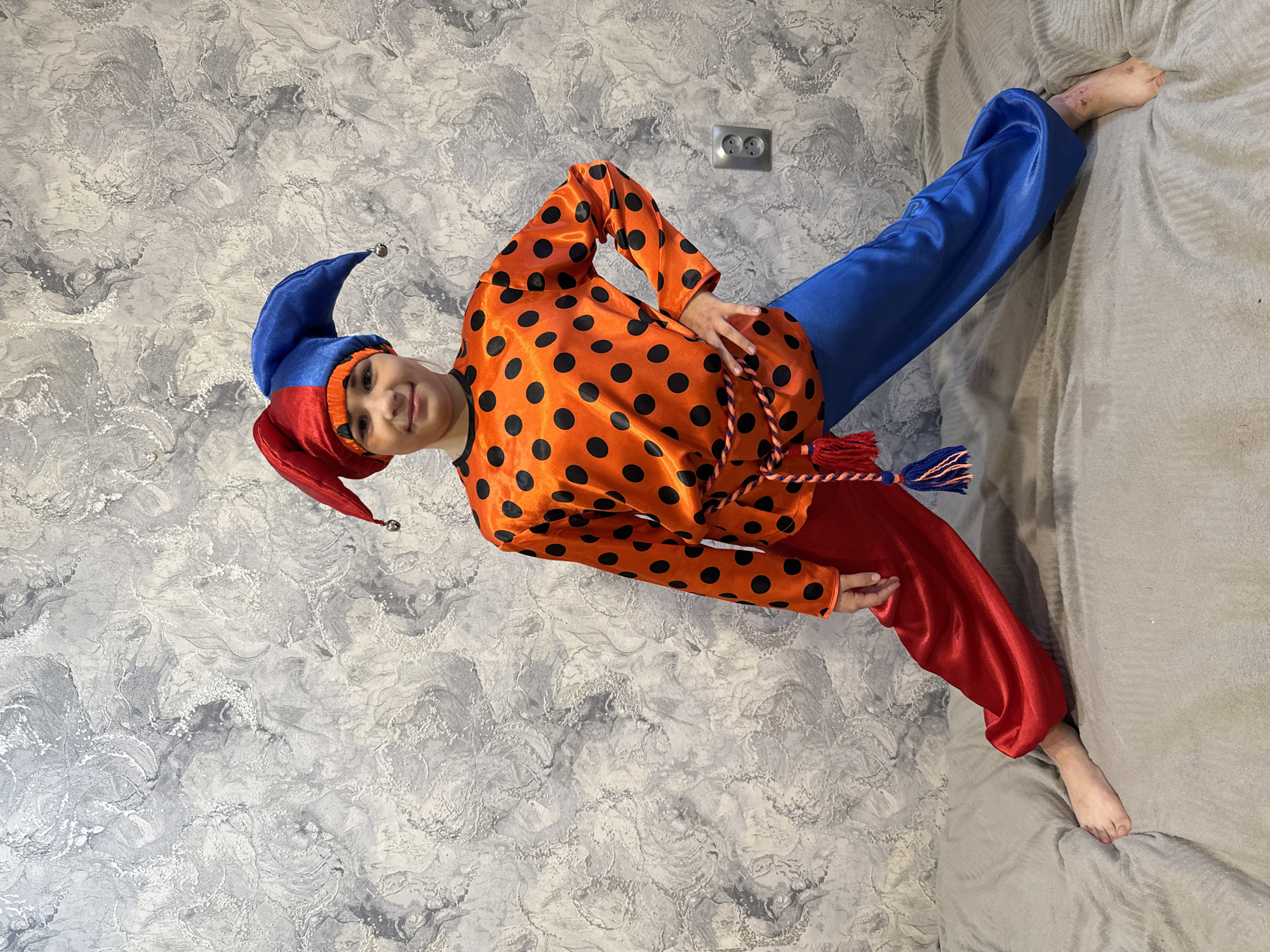 An indoor, full-length portrait captures a child dressed in a vibrant jester or clown costume, posing against a textured gray and white wallpapered wall in Rusanovo, Russia. The child, smiling slightly, stands with one hand on their hip, looking directly at the camera.
The colorful costume features a blue and red jester hat adorned with a small bell at the tip of its blue side. They wear a long-sleeved, bright orange top covered in large black polka dots. Their pants are distinctly mismatched, with one leg blue and the other red. A multi-colored rope, featuring red, blue, and orange tassels, is tied around the child's waist.
The background consists of a light gray wall with a swirled or marbled texture. An electrical outlet is visible on the wall to the right of the child's head. To the lower right, a soft, light gray upholstered surface, likely a bed or sofa, is present. The child's bare feet are visible, one resting on this surface. The lighting suggests an indoor setting, likely with ambient natural or artificial light. There is no visible text in the image.