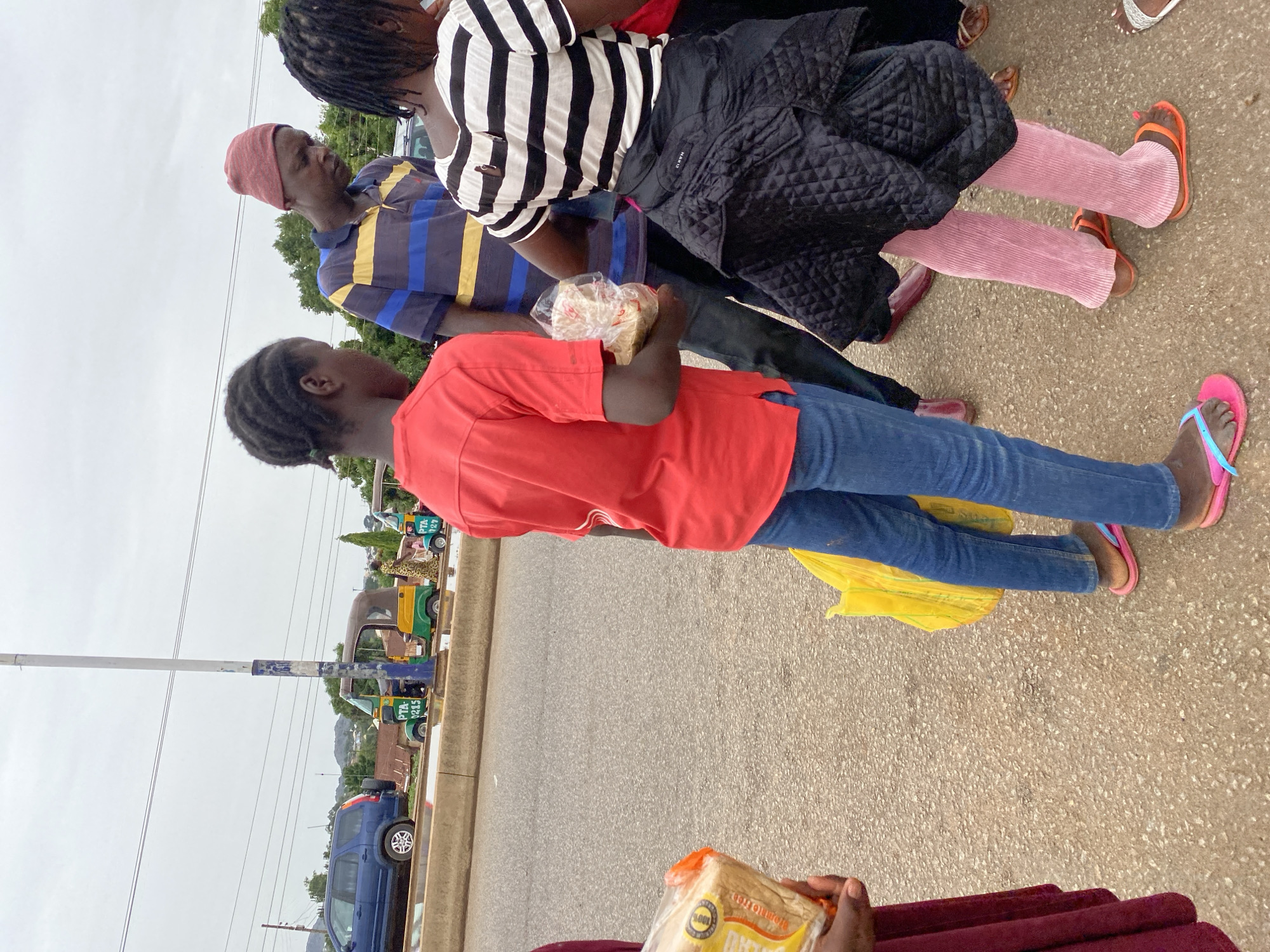 The image shows a group of people standing on the side of a road. One person in the foreground is holding a loaf of sliced bread. Several cars, a few "keke" auto-rickshaws, and a pole are visible on the side of the road in the background. The location is Bukuru, Nigeria.