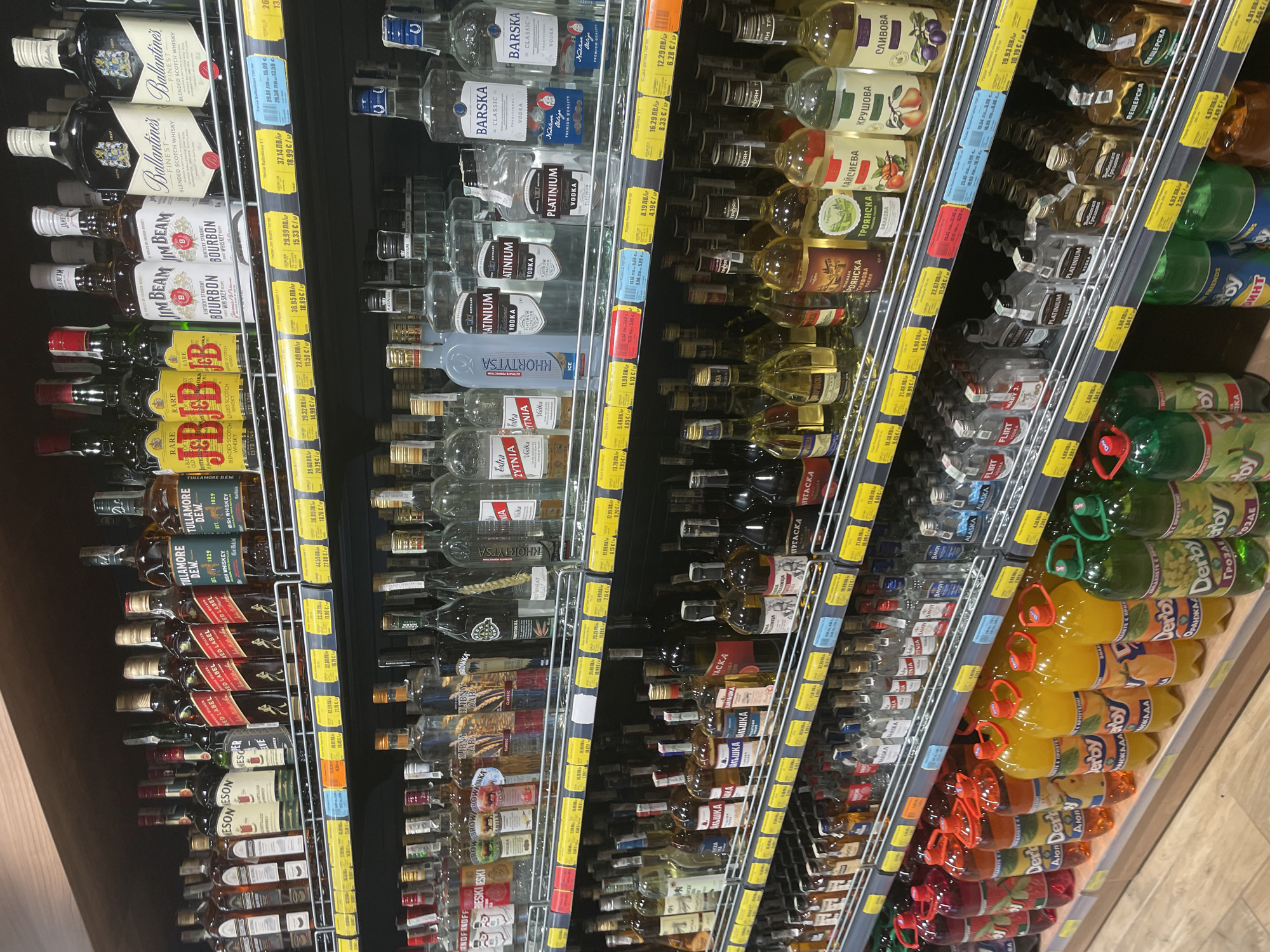 The image depicts a well-lit interior aisle of a supermarket in Ihtiman, Bulgaria, specifically showcasing a section dedicated to alcoholic beverages. Multiple metal-frame shelves are fully stocked with various bottles of spirits, presented for retail sale. No individuals are present within the frame.
The top shelf displays numerous whiskey and scotch brands, including multiple bottles of Johnnie Walker Red Label, Tullamore D.E.W. Irish Whiskey, Jameson Irish Whiskey, J&B Rare Scotch Whisky, Jim Beam Bourbon, and Ballantine's Finest Blended Scotch Whisky. Each product is accompanied by a yellow pricing label indicating prices in both Bulgarian Leva (ЛВ) and Euros (€). Examples of pricing include 77.80 ЛВ / 39.79 € for Tullamore D.E.W., and other prices ranging from approximately 22.49 ЛВ / 11.50 € to 39.68 ЛВ / 20.29 €.
The second shelf from the top features a diverse selection of vodkas and other spirits. Identifiable brands include Smirnoff, Khortytsa Vodka, Platinum Vodka, and Barska Classic Vodka. Pricing labels are consistent with the top shelf, showing prices such as 13.05 ЛВ / 6.67 € for Khortytsa Ice, 11.19 ЛВ / 5.72 € for Platinum Vodka, and 16.29 ЛВ / 8.33 € for Barska Classic.
The third shelf also contains a variety of vodkas, brandies, and fruit-based spirits, including bottles labeled 'Крушова' (Pear), 'Сливова' (Plum), and 'Кайсиева' (Apricot). Other brands like Trojanska and Burgaska are visible. Prices on this shelf range from approximately 16.98 ЛВ / 8.68 € to 23.49 ЛВ / 11.99 €.
The bottom shelf presents a combination of smaller, miniature bottles of spirits (e.g., Flirt Vodka, Mary J, Platinum, Ябълкова градина, Нещерска) and large plastic bottles of non-alcoholic beverages. The large bottles are from the "Derby" brand, appearing in various fruit flavors (red, orange, yellow, green), likely sodas or juices. Miniature bottle prices include 4.68 ЛВ / 2.39 € and 2.82 ЛВ / 1.44 €. Derby drinks are priced around 4.67 ЛВ / 2.39 €.
The shelving units are constructed from dark metal with wire racks. The background wall is dark, contrasting with the illuminated products. The floor exhibits a light wood-grain pattern. Overhead fluorescent lighting provides uniform illumination throughout the aisle.