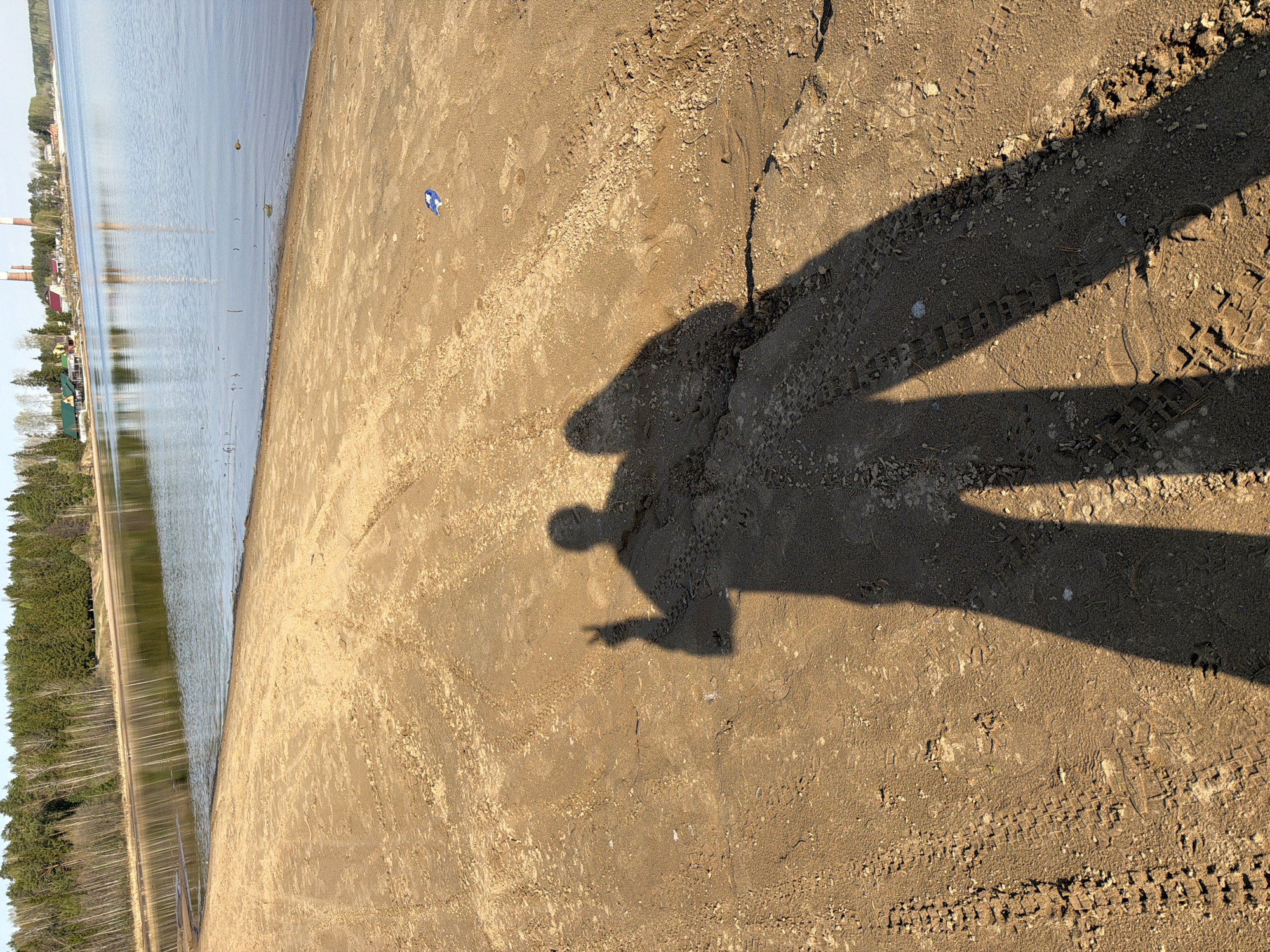 The image depicts a sandy beach in Chaikovsky, Russia, adjacent to a body of water. The water is lined with trees and distant buildings. A human shadow is cast on the sand, showing a person with one arm raised. Tire tracks are visible in the sand. A piece of blue and white debris is located nearby.