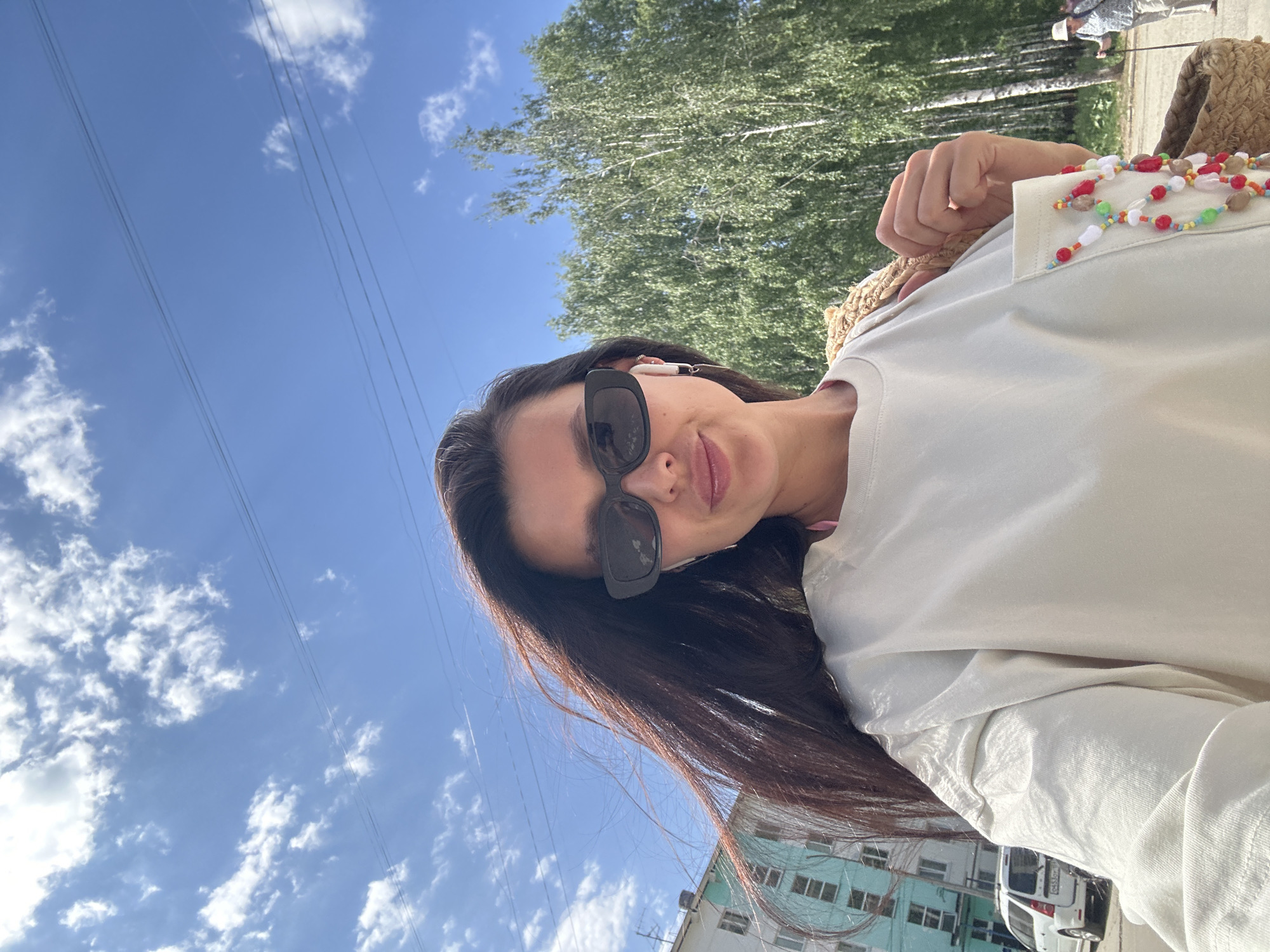 A woman with long dark hair and sunglasses poses for a selfie. She is wearing a light-colored shirt and a straw bag with a beaded necklace is visible. Behind her is a blue sky with scattered clouds and a stand of birch trees. Power lines stretch across the sky. In the background, a multi-story building painted in light blue and white can be seen, along with a white van parked near the building. The image was likely taken in Chaikovsky, Russia, given the location request.