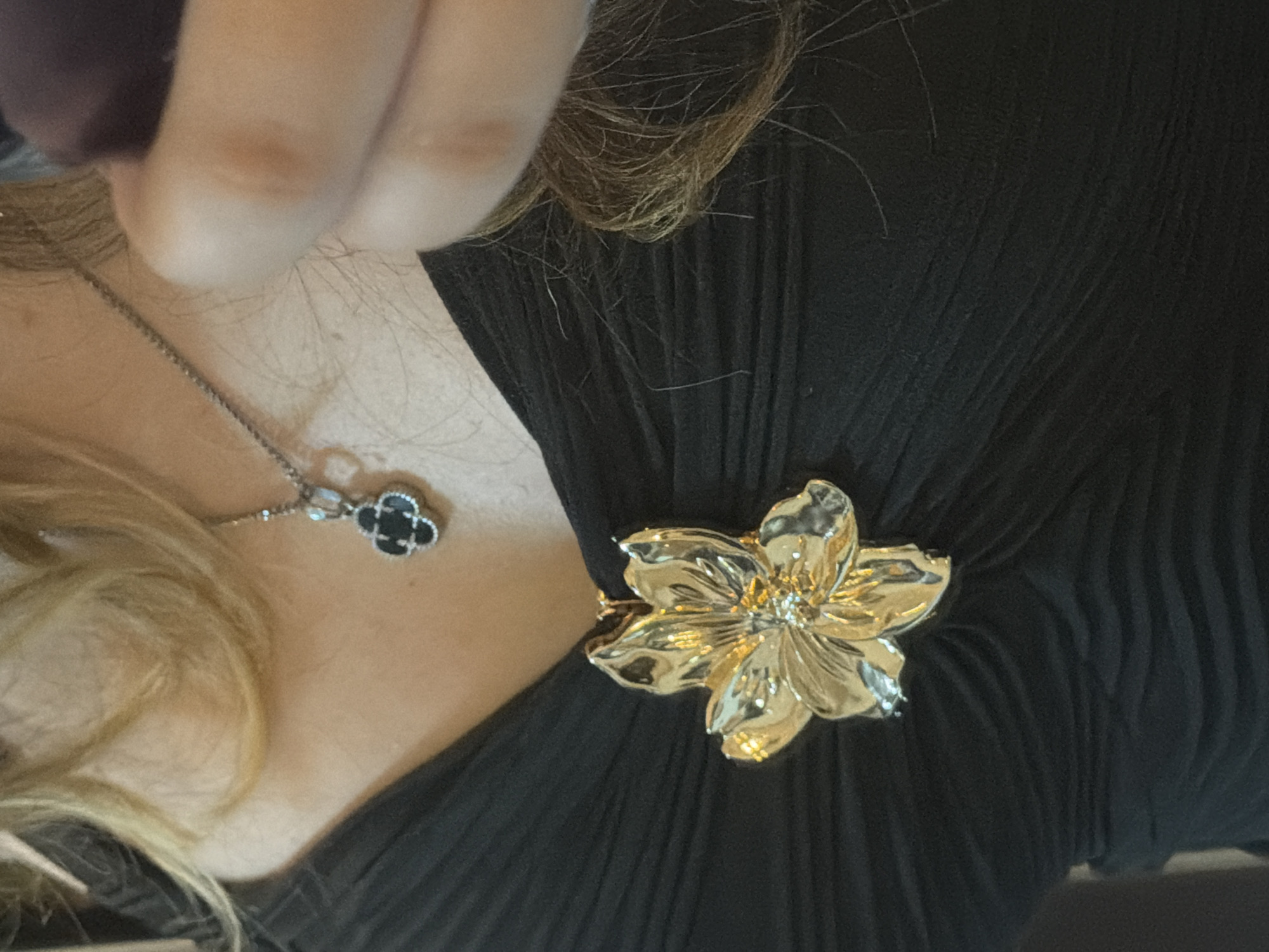 The image shows a close-up of a person's chest area, adorned with jewelry. A prominent gold flower-shaped brooch is pinned to a black, ruched garment. A delicate silver chain with a clover-shaped pendant, featuring black stones, hangs around the person's neck. The person's fair hair is visible around the neck and shoulders, and a blurred hand holding a dark object is in the upper right corner of the frame. The background is dark and indistinct, suggesting an indoor setting. The exact location in Plovdiv, Bulgaria, cannot be determined from the provided image.