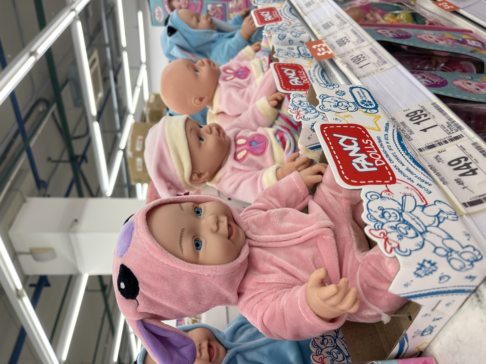 The image shows a shelf of "Fancy Dolls" for sale in what appears to be a store. The dolls are in boxes and are dressed in pink and blue bunny-themed outfits. Price tags are visible, with a price of 449 руб (Rubles). Text in Russian indicates the item is "КУКЛА FANCY DOLLD ЛЫШ СТИЧИК," or "Doll Fancy Doll Stitch." The text also indicates the product is made in Belarus. Other prices visible are 199 руб and 1199 руб. The location is indicated by the text as Izhevsk, Russia. The dolls are lined up on the shelf and are the primary focus of the image.