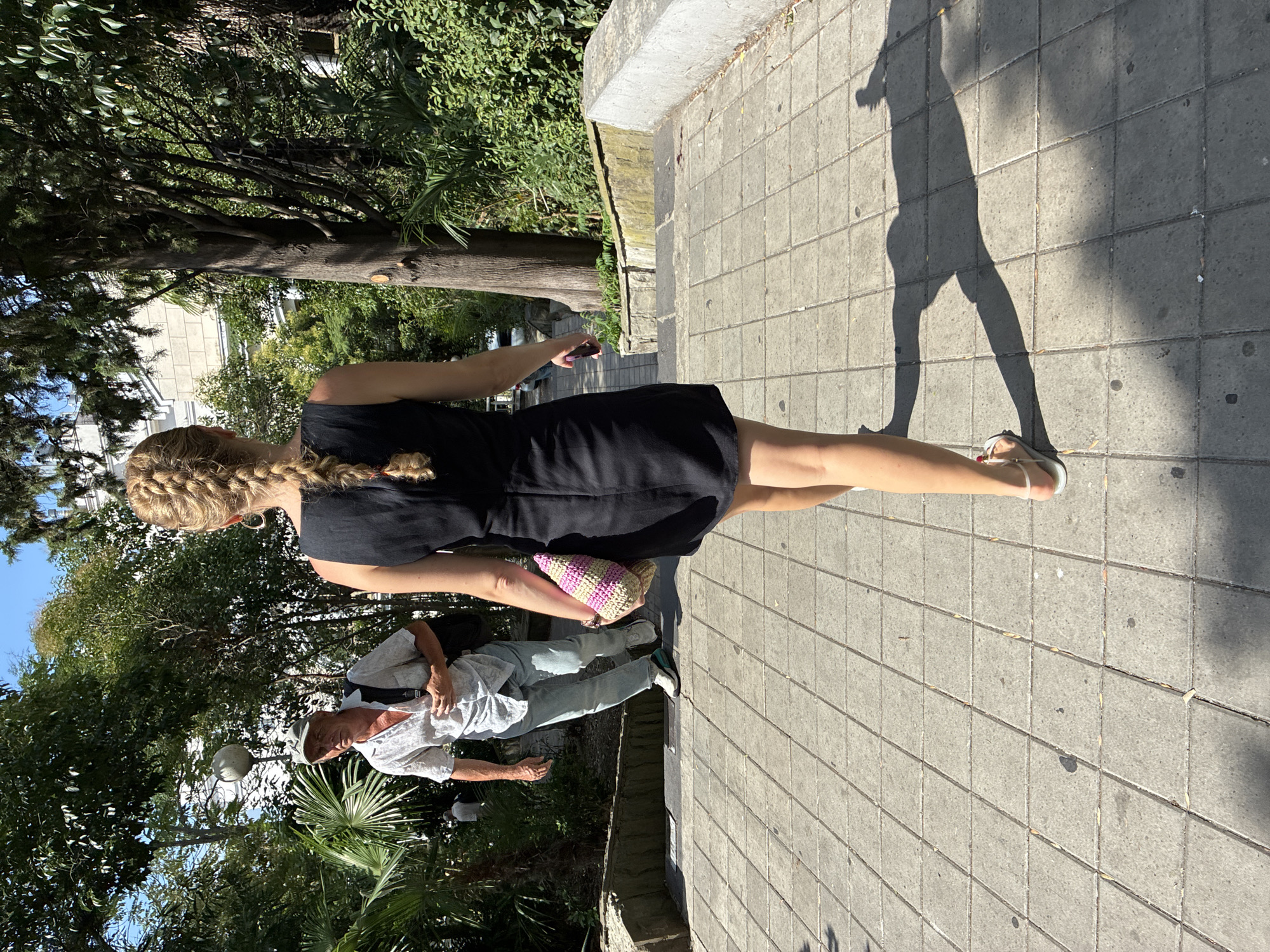 In Sochi, Russia, on a sunny day, a woman with blonde hair in a braid walks away from the camera on a tiled pathway lined with greenery. She wears a black sleeveless dress and light-colored sandals, carrying a woven bag. To her left, further up the path, a man in a light-colored shirt, blue jeans, a cap, and white sneakers walks down the stairs. He is carrying a backpack. The environment includes lush trees, palm trees, and a building in the background.