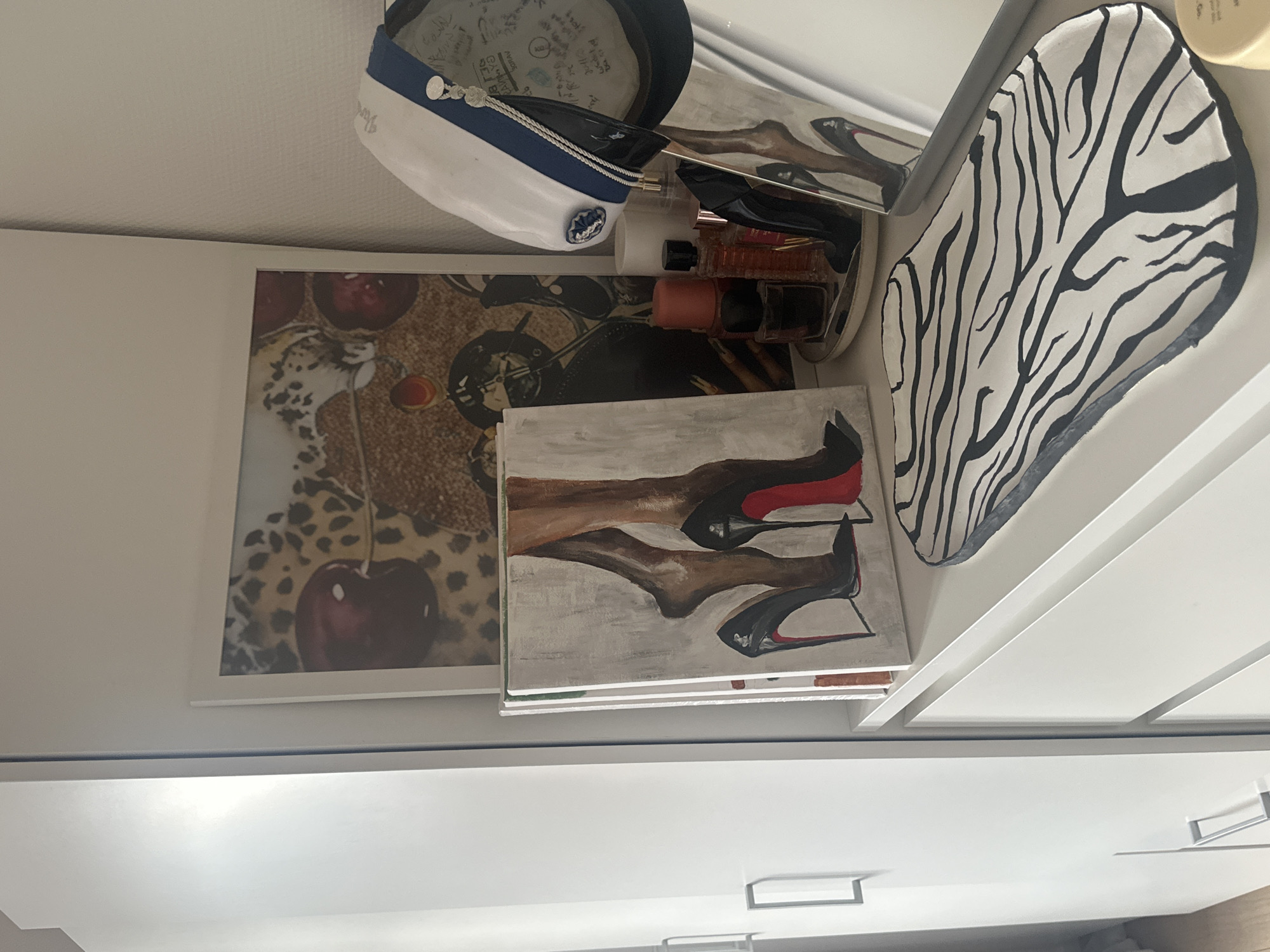 The image shows a dresser with several decorative items on top. There is a framed artwork of cherries and a leopard print, a painting of a woman's legs in high heels, several perfume bottles on a tray, and a decorative tray with a zebra print pattern. A white hat with a blue and gold trim is also visible. The dresser is white and has silver handles. The background shows a white wall. The scene is indoors, likely a bedroom or dressing area. The location is identified as Varde, Denmark. There are no people present in the image.