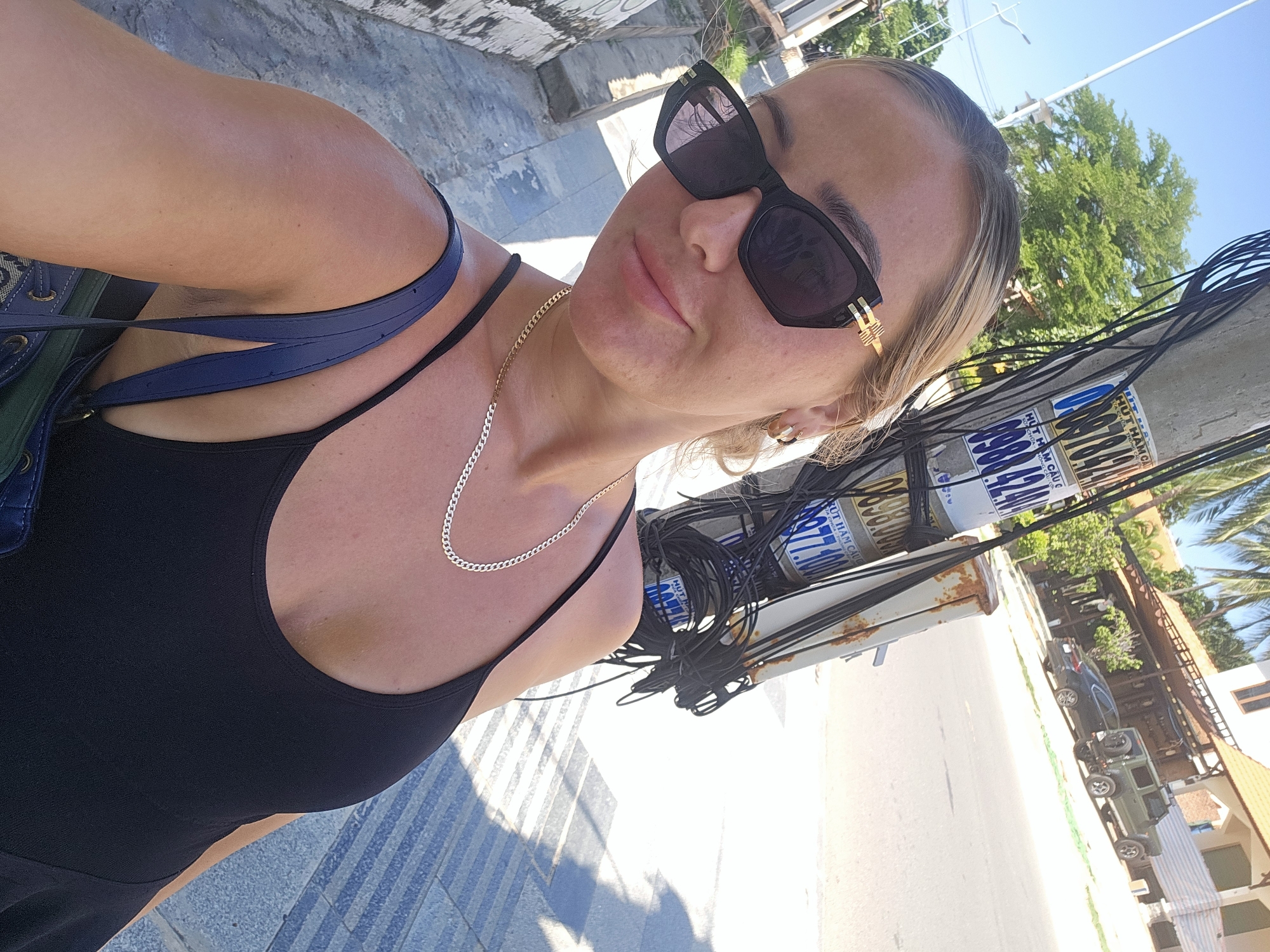 A woman is taking a selfie outdoors on a sunny day in Phu Thuy Ward, Vietnam. She is wearing sunglasses, a black tank top, and a gold chain necklace. Behind her, a utility pole with many wires is visible, along with a street and buildings. The ground is paved with gray and blue tiles.
