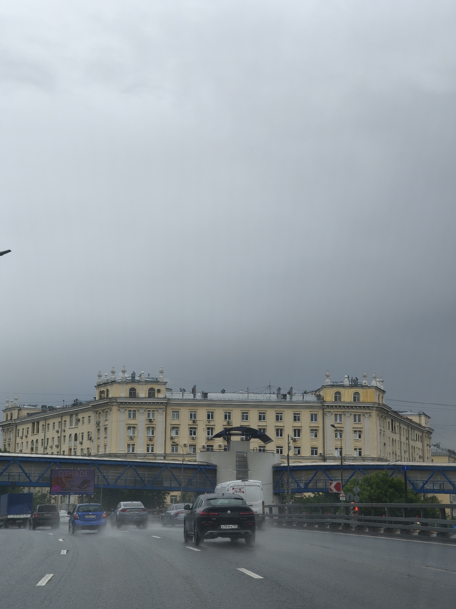 The image depicts a street scene in Moscow, Russia, during a rainy day. The road is wet and several vehicles, including cars and a van, are driving on it. A large, light yellow building with decorative elements is situated in the background. Pedestrian overpasses, one of which has an advertisement on it, are visible in the mid-ground. The sky is overcast and grey, indicating the rainy weather conditions.