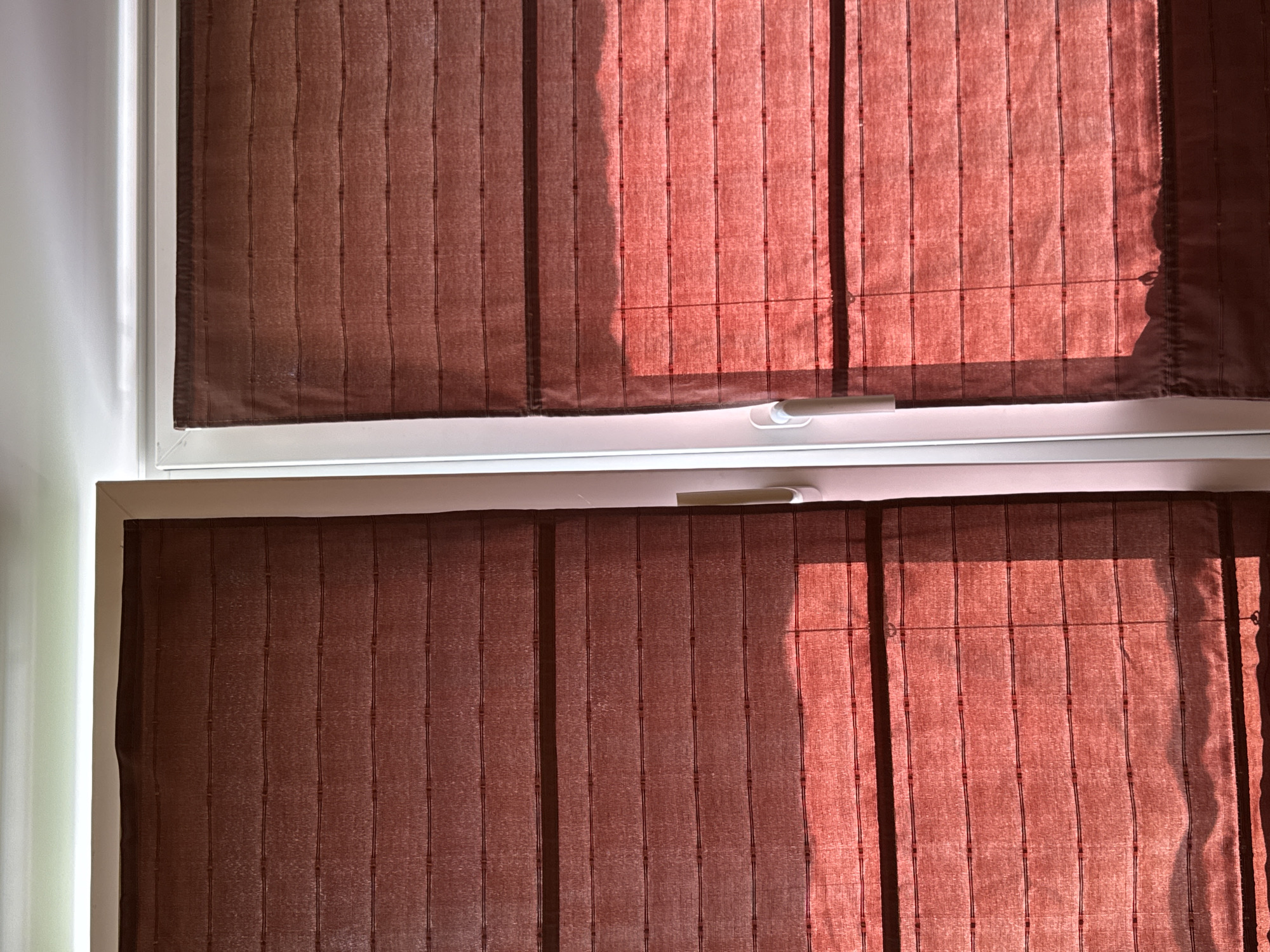 This is a close-up, eye-level, indoor shot of a window with two red, textured roman blinds. The blinds are partially lowered, with the top sections obscured by the window frame. Horizontal seams and dark, thin stitching are visible across the fabric of the blinds, creating a striped effect. Sunlight streams through the blinds, casting a warm red glow and creating dappled shadows on the fabric, particularly on the right-hand blind. The window frame is white, and a thin white ceiling or trim is visible at the top of the image. A white handle is attached to the right-hand side of the window frame. The scene appears to be in a residential setting, possibly a bedroom or living room, in the city of Yarominski Sel'ski Savet, Belarus. There are no people visible in the image.
