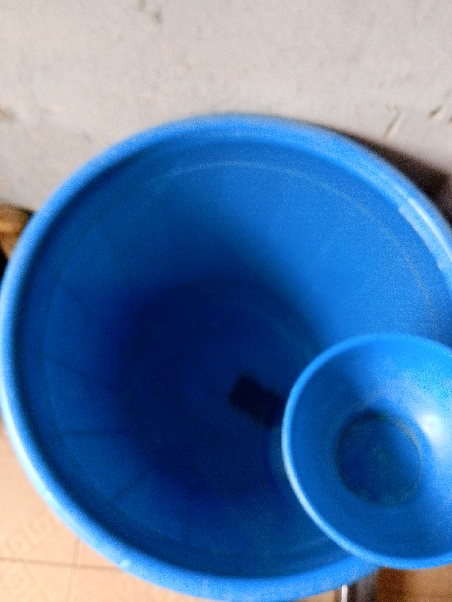 In an indoor setting, two blue plastic containers are visible. The larger container is a wide, cylindrical bucket. Nestled inside the larger bucket is a smaller, conical container. The background shows a textured, light-colored wall and a glimpse of a tiled floor. The lighting appears to be natural, casting shadows on the objects. The image is slightly out of focus.