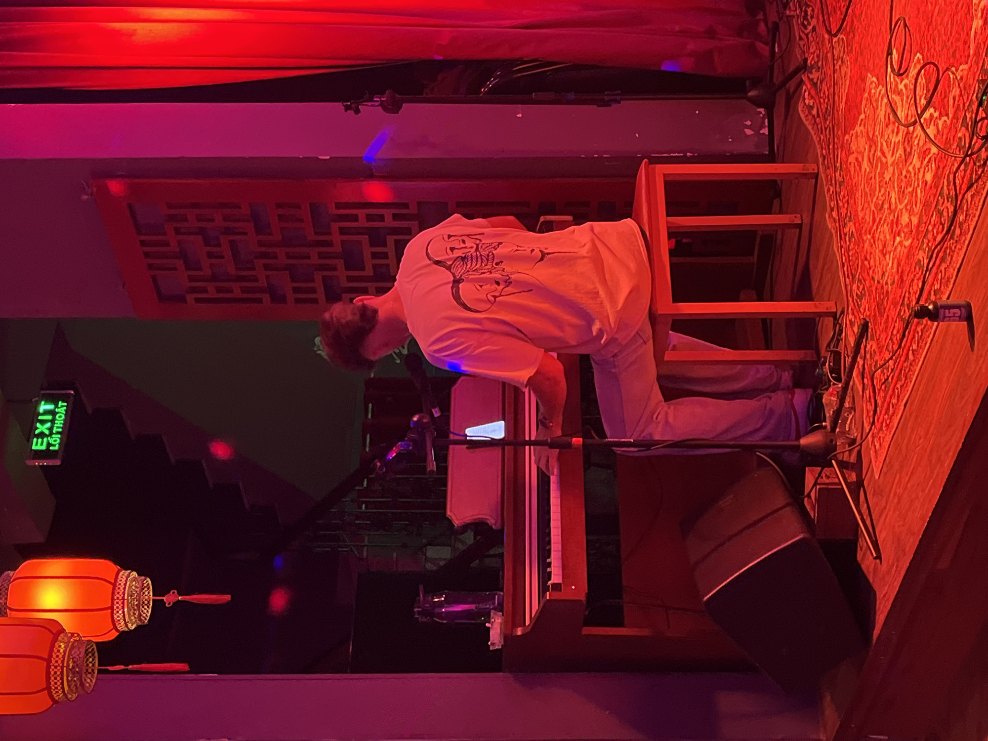 A person is playing an organ on a stage in a room, likely in Đà Nẵng, Vietnam. The person wears a white t-shirt with a print on the back and light-colored pants. They are seated on a wooden chair in front of the organ. A microphone stand stands in front of the organ. A large speaker is beside the organ. Overhead, there is red stage lighting and red decorative panels. To the left, there are illuminated red paper lanterns and an exit sign in Vietnamese. In the background, there are stairs. To the right of the organ, there is a red patterned rug on the floor and other stage equipment including microphone stands and cables.