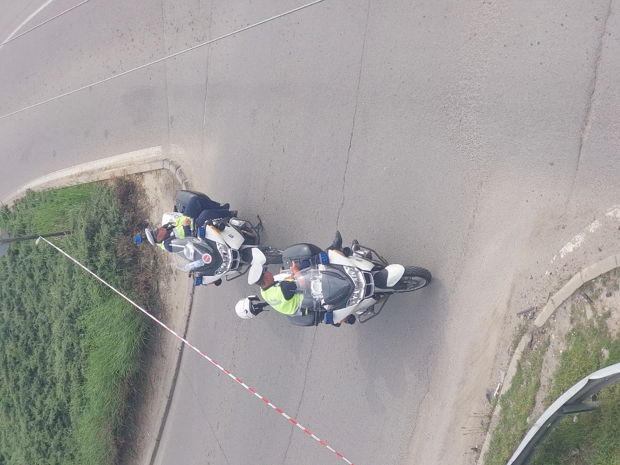 Two uniformed police officers on motorcycles are positioned on the side of a gray asphalt road in Sofia, Bulgaria. One officer is wearing a white helmet and a yellow vest, while the other is wearing a blue helmet and a yellow vest. A red and white striped barrier tape runs along the edge of a grassy area adjacent to the road. The area includes a concrete curb. A metal guardrail is visible in the bottom right corner.
