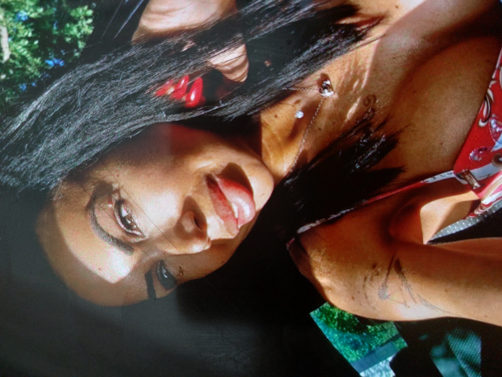 The image is a close-up shot of a woman with dark hair lying down. Her face is tilted towards the camera, with her tongue slightly sticking out. She has a necklace with a pendant. A portion of her shoulder and arm is visible, revealing a tattoo. She's wearing a red and white top. The background is blurred, with glimpses of greenery suggesting an outdoor setting. There is no information about the location in the city of Tsarevo, Bulgaria.