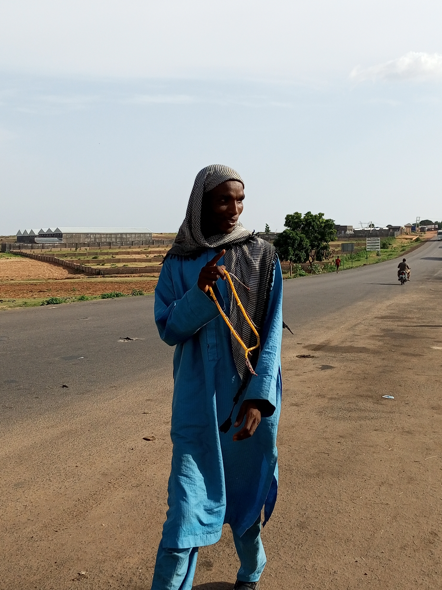 A man in a blue robe and head covering walks on the side of a road in Gadao, Nigeria. He is holding a string of yellow beads in his right hand. The road is paved and appears to be a main thoroughfare, with a few vehicles and pedestrians visible in the distance. The landscape around the road is dry and dusty, with some sparse vegetation and agricultural fields. In the background, there are several large, modern buildings that appear to be industrial or agricultural structures. The sky is clear and blue with a few scattered clouds. The overall scene suggests a rural or semi-rural setting in Nigeria.