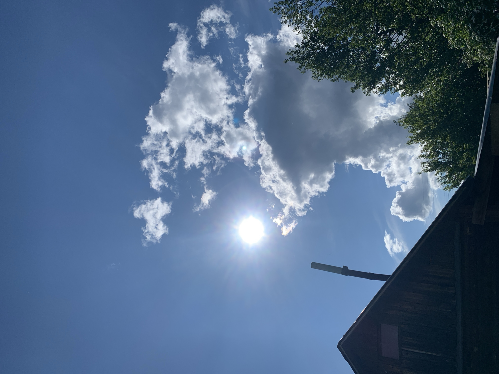 The image shows a daytime scene with a clear blue sky and scattered white clouds. The sun is visible, shining brightly and creating lens flare. In the upper right corner, there are green tree leaves. A portion of a dark, weathered wooden building is visible in the lower right corner, with a pipe extending upwards from the roof. The location, Chaikovsky, Russia, cannot be confirmed based on the image content alone.