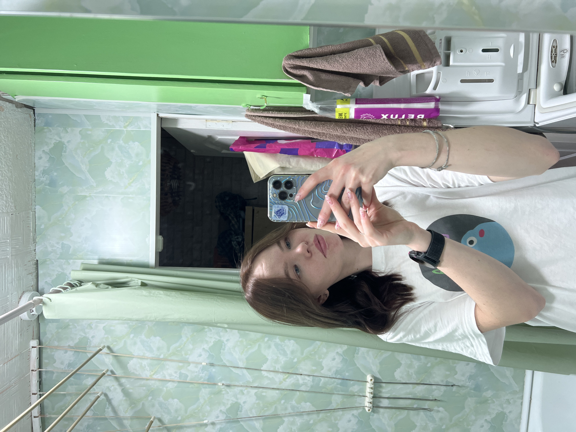 A young woman is captured in a mirror selfie within what appears to be a bathroom or utility room in Chaikovsky, Russia. She has long, dark brown hair and light eyes, with a subtle natural makeup look. She wears a plain white t-shirt featuring a graphic design of a blue, cartoon-like character with a pink nose on a black circular background. On her left wrist, she wears a black smartwatch, and on her right wrist, two silver bracelets. Her manicured nails, painted in a light pink or nude color with intricate white designs, are visible as she holds a smartphone. The phone's case is distinctive, featuring a wavy, topographic-like pattern in blue and silver.
The background, reflected in the mirror, reveals elements of the room. A green wall or cabinet is visible at the top, and the walls below it are covered in light green and white marbled-patterned tiles or wallpaper. To the right, a white washing machine is partially visible, specifically its open detergent dispenser tray with universal symbols for washing cycles. Brown towels, one with stripes, are draped nearby. A purple package, possibly containing feminine hygiene products, is also present, displaying reversed Cyrillic text that appears to read "БЕГЛЯ" (BEGLYA). A pink and purple plastic bag is tucked underneath. To the left, a multi-tiered clothes drying rack with white plastic frames and metal rods is set against the marbled wall. A light green shower curtain or fabric screen hangs to the left, and a reflection of a darker, possibly unfinished wall surface is seen further back. The overall lighting suggests indoor artificial light. The woman's expression is calm and neutral.