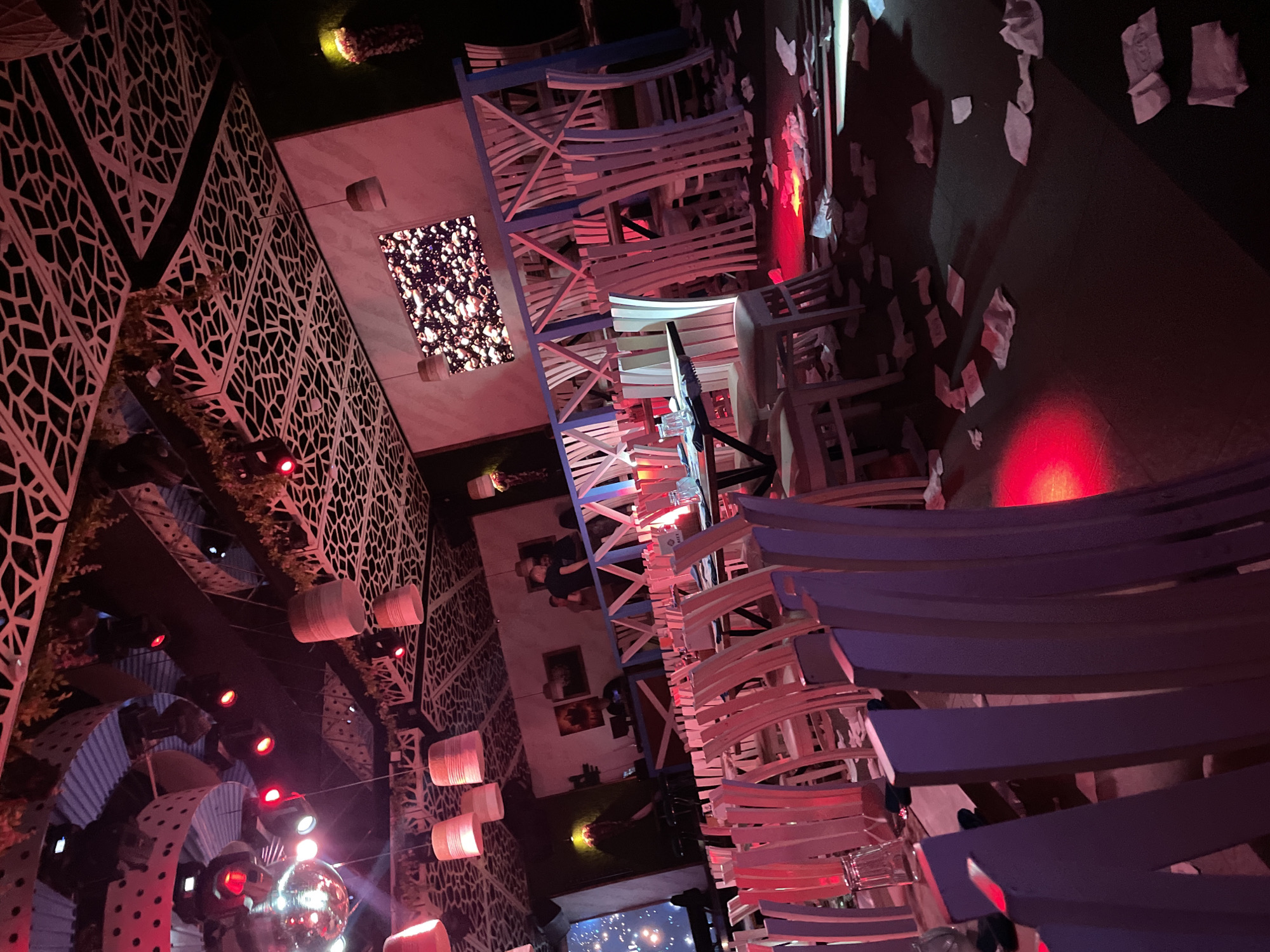 The image shows a brightly lit, elaborately decorated interior space, potentially a restaurant or event venue, in Plovdiv, Bulgaria. The scene includes a multi-layered structure built with white and blue chairs. The ceiling is draped with white papers and illuminated by red and white lighting. A large disco ball and several decorative lanterns are also visible. There are lattice-patterned walls and some potted plants along with multiple people in the background.