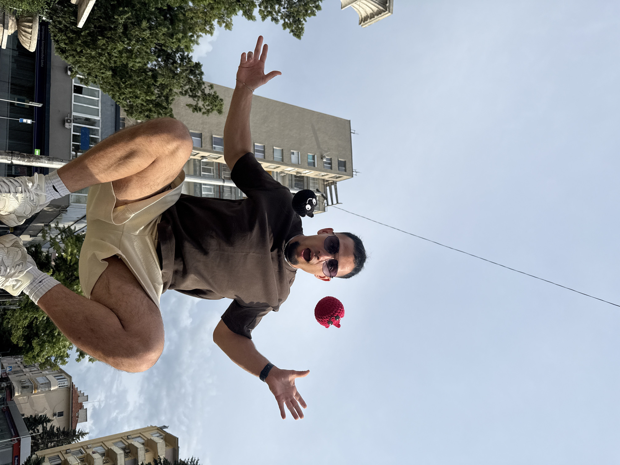 The image, taken from a low angle, shows a man in mid-air, appearing to be jumping or falling. He is wearing a brown t-shirt, light-colored shorts, white sneakers, and sunglasses. His mouth is open in surprise or excitement, and his arms are outstretched. In front of him, suspended in the air, are two small crocheted objects: one is a black creature with large white eyes, and the other is a red, octopus-like figure with red eyes.
The background consists of a pale blue sky with scattered clouds, suggesting it is daytime with fair weather. Buildings are visible at the top and bottom of the frame, with trees peeking out from behind them. One building, a multi-story structure, appears to be an apartment building. The architecture suggests an urban environment.
The man's posture and facial expression, combined with the floating objects, create a whimsical and dynamic scene. The perspective, looking up at the subject against the sky, emphasizes the sense of movement and height. No visible text is present in the image. The scene is set in Shumen, Bulgaria.