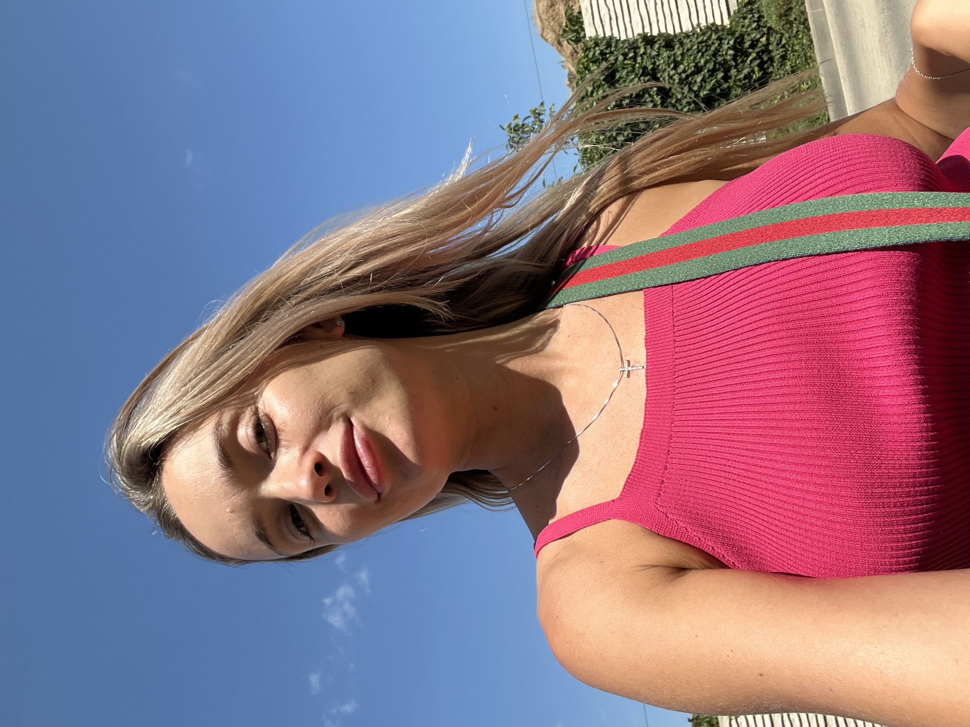 A young blonde woman is captured in a low-angle, close-up selfie against a bright blue sky with scattered clouds. She is wearing a bright pink, ribbed tank top and a silver necklace with a cross pendant. A strap with green and red stripes, possibly from a bag, is visible crossing her chest. Her hair is blowing gently in the wind. In the background, there is a white fence covered in green ivy and a hint of a building and hillside. The overall scene suggests an outdoor setting, likely in a suburban or rural area. The location is identified as Kryzhanivka, Ukraine.