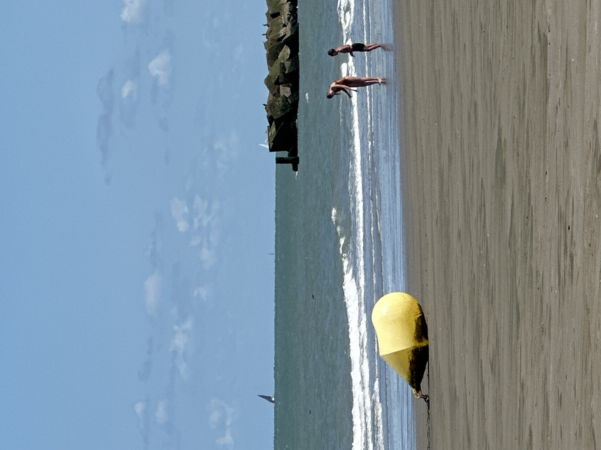 A wide shot of a sandy beach in Dunkirk, France, shows the sea, a breakwater, and a yellow buoy. The water is a deep green, with white waves crashing near the shore. Two people, an adult and a child, stand in the shallows. The adult appears to be bending down. The breakwater is made of large, rough-hewn rocks and extends out into the sea. A sailboat is visible on the horizon. The sky is a clear blue with scattered white clouds. The yellow buoy is partially submerged in the sand near the foreground.