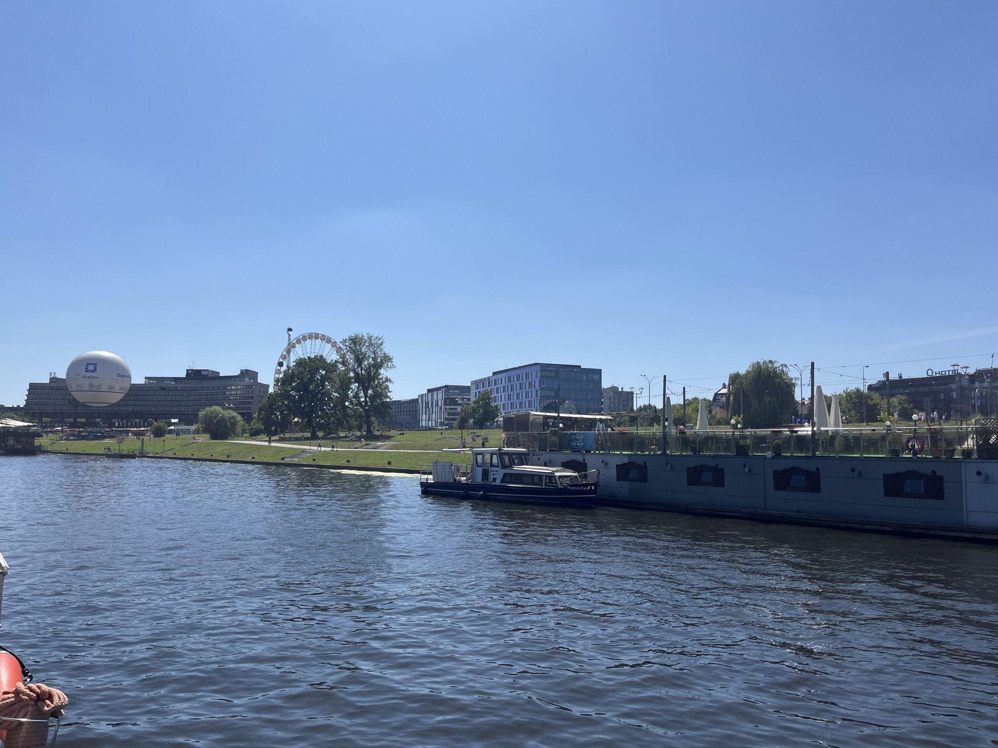 This image captures a sunny day in Krakow, Poland, along the Vistula River. In the foreground, a calm, dark blue river flows, reflecting the clear sky. A boat, partially visible on the left, has a red life preserver and rope. In the midground, a long, gray boat is docked along the riverbank, its side decorated with square panels. A smaller blue and white boat is moored alongside it. The riverbank is lined with a green lawn, and a pathway is visible. Beyond the embankment, a large Ferris wheel stands tall, and a white blimp with "Dobra Energia" and "Kraków" written on it is visible to the left of the Ferris wheel. Several modern buildings with glass facades are situated behind the green space, including one with "CAMPUS" written on it and another with "Q HOTEL". Trees are scattered throughout the scene, and a few people are visible walking along the pathway. The sky is a bright, cloudless blue.